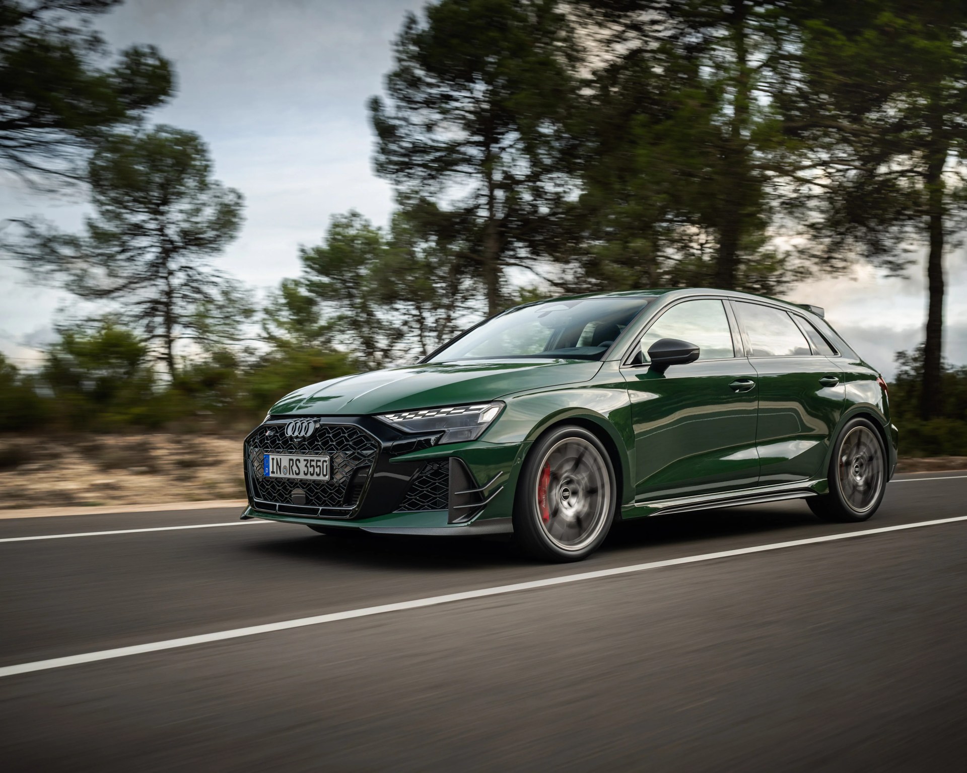 Dark green Audi RS3 Sportback driving on a road with blurred wheels and trees in the background.