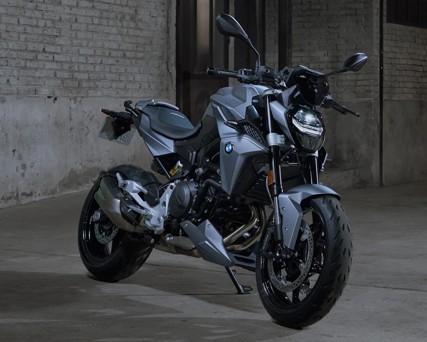 Matte gray BMW motorcycle with black accents and dual disc brakes parked indoors against a brick wall.