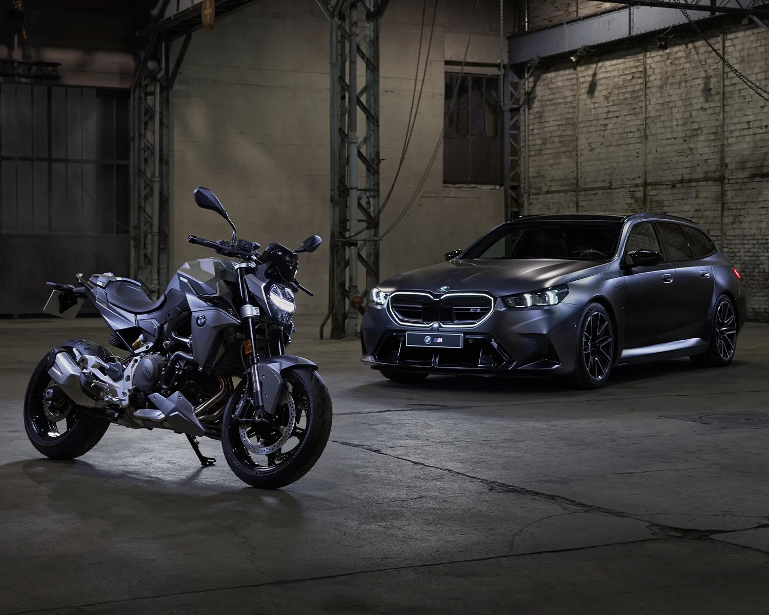 Matte gray BMW motorcycle and BMW station wagon parked inside an industrial warehouse.