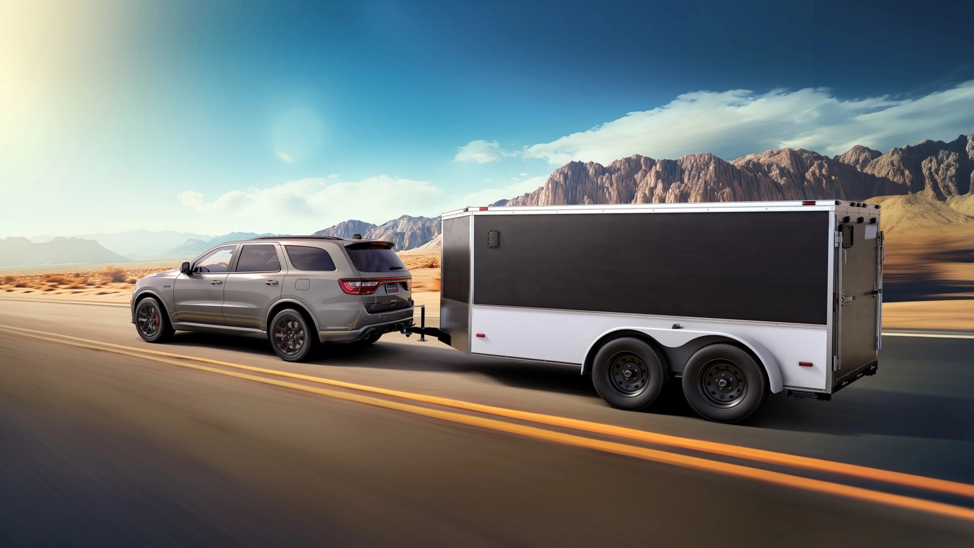 Gray SUV towing a black and white enclosed trailer on a desert highway with mountains in the background.