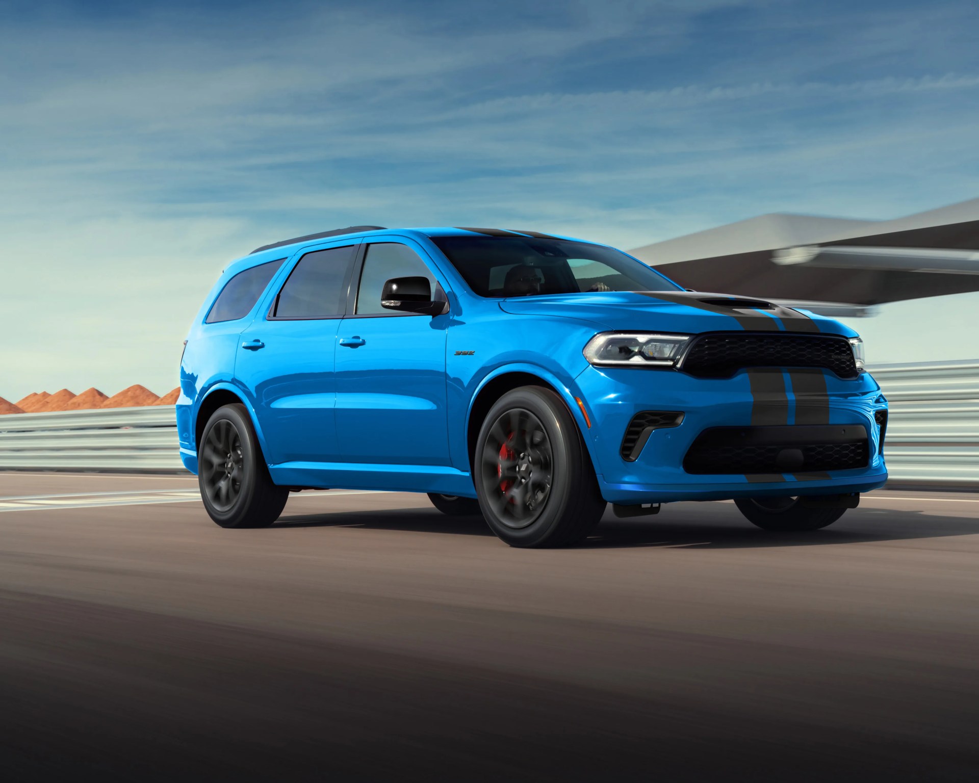 Blue Dodge Durango SUV with black racing stripes driving on a racetrack.