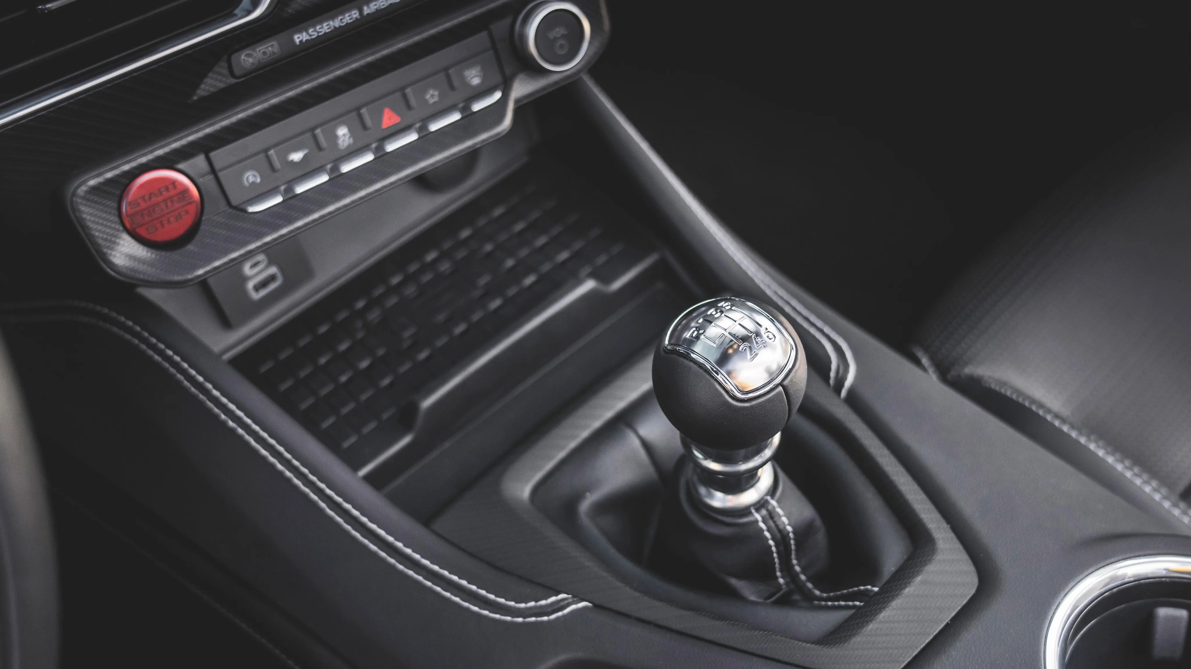 Black leather manual gear shift knob with silver top and white stitching in a car interior.