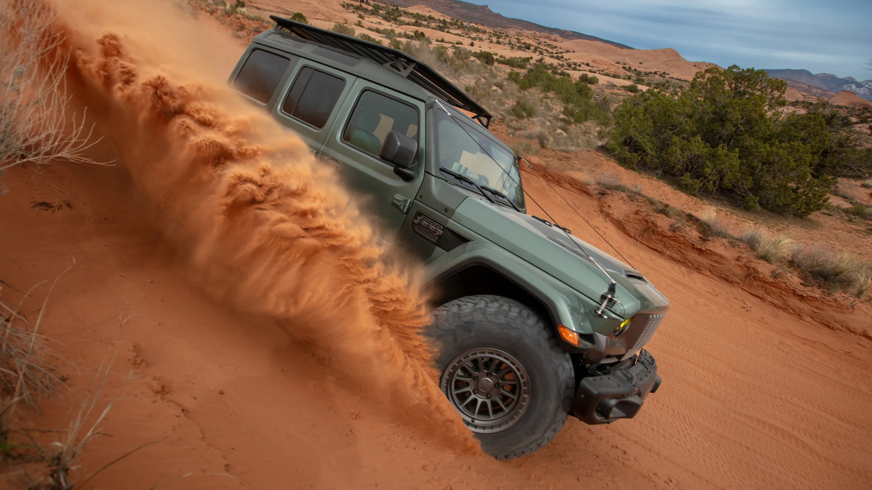Green off-road SUV kicking up red desert sand while driving on a sandy trail with bushes and rocky hills in the background.