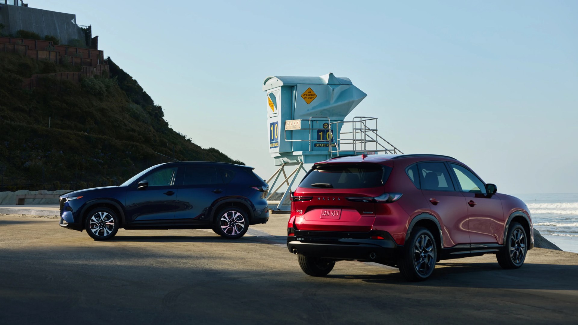 Two Mazda SUVs, one dark blue and one red, parked near a blue lifeguard tower by the beach.