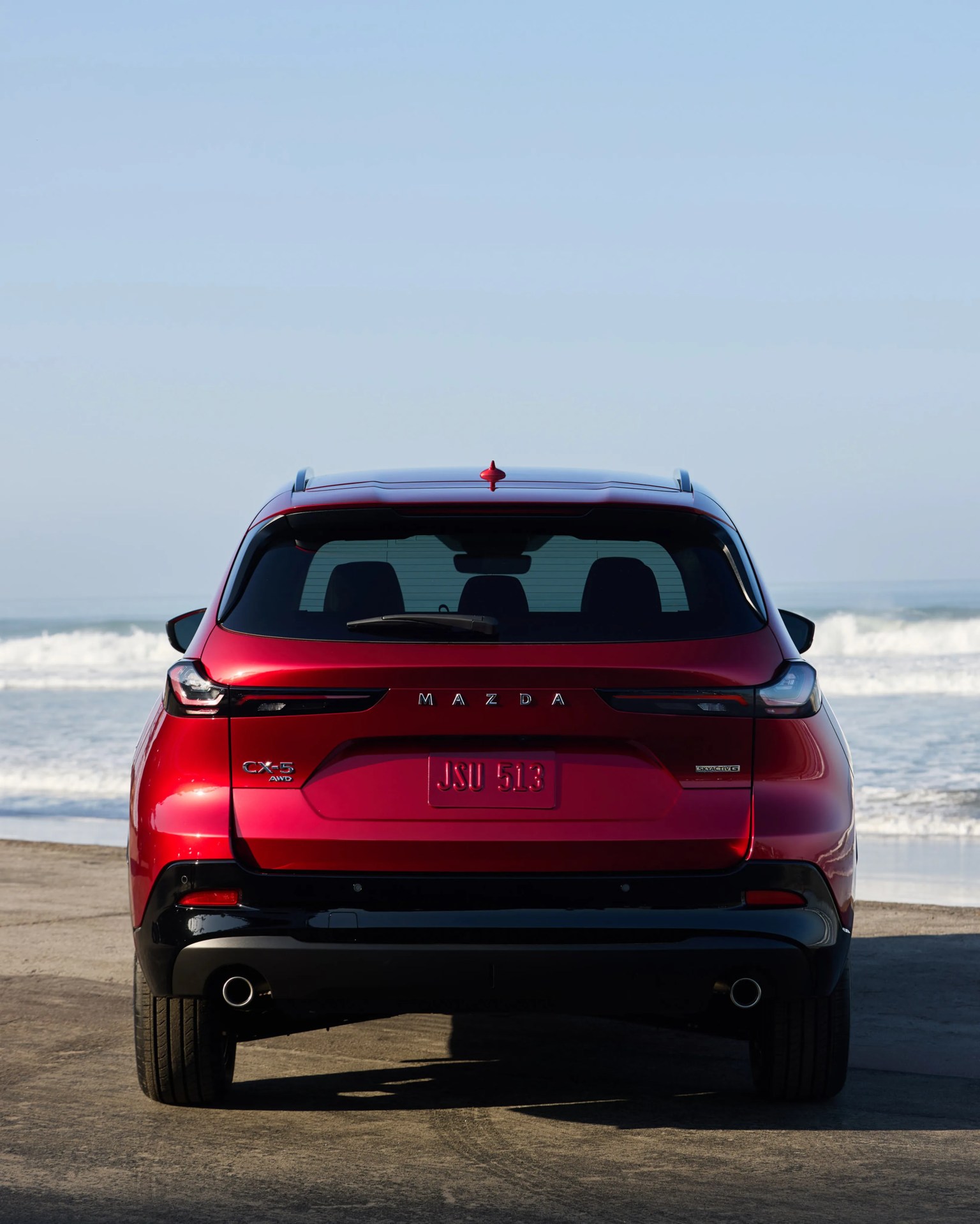 Rear view of a red Mazda CX-5 AWD SUV parked on a beach with ocean waves in the background.