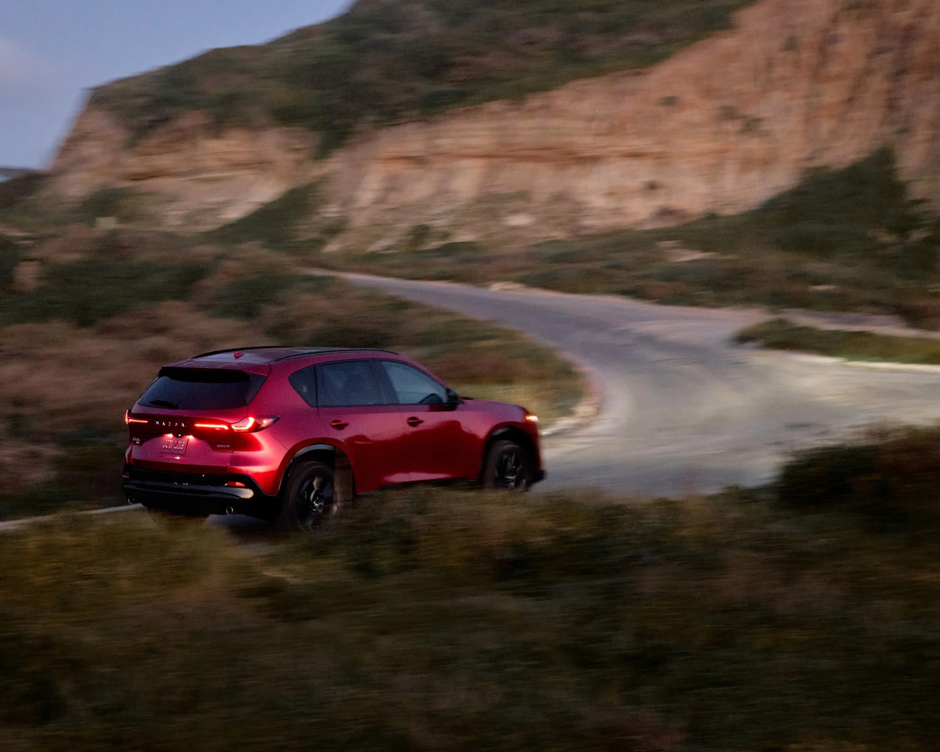 Red Mazda SUV with illuminated rear lights driving on a winding road near rocky hills at dusk.