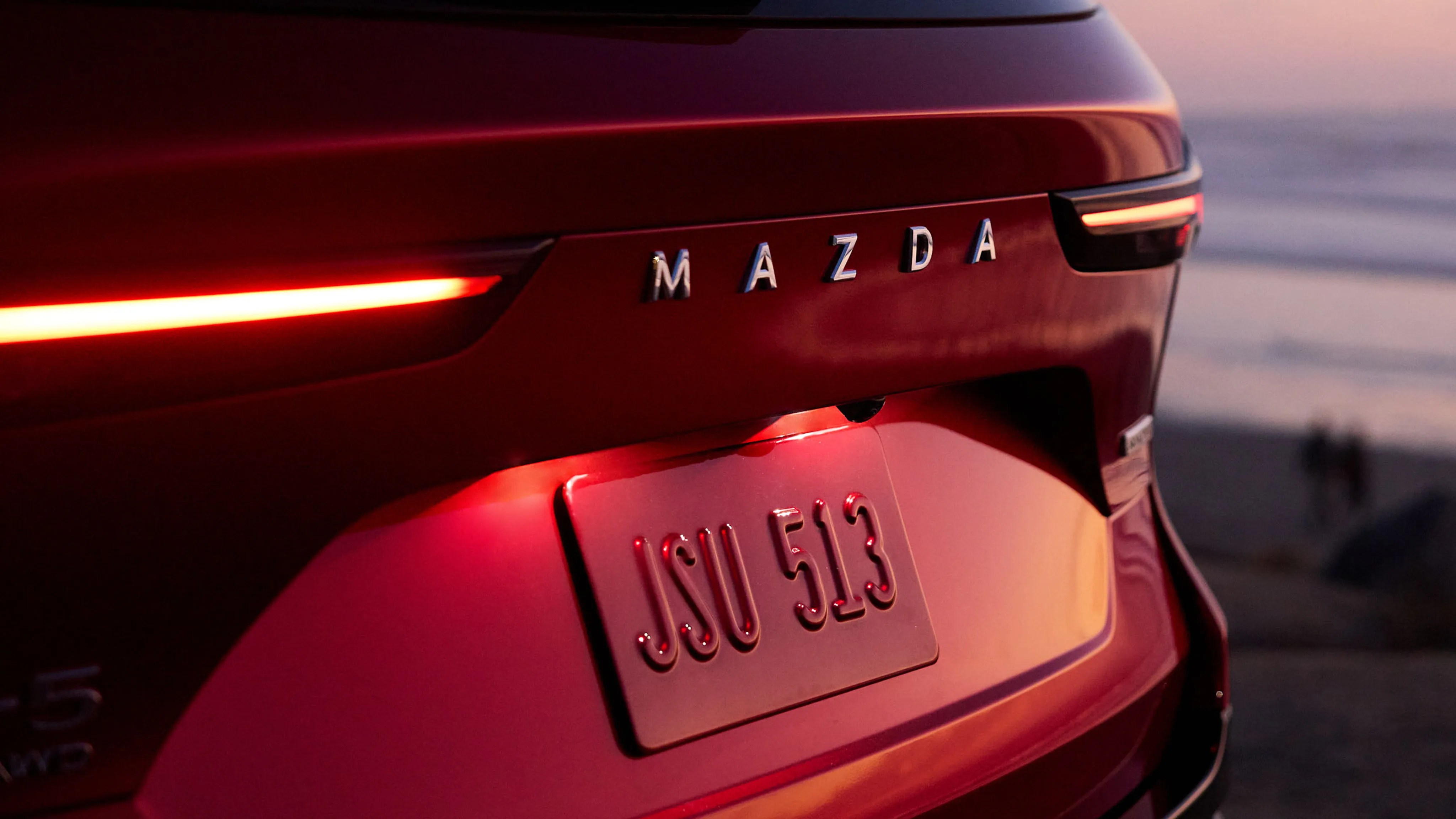Rear view of a red Mazda car with illuminated taillights and a license plate reading "JSU 513" at sunset.