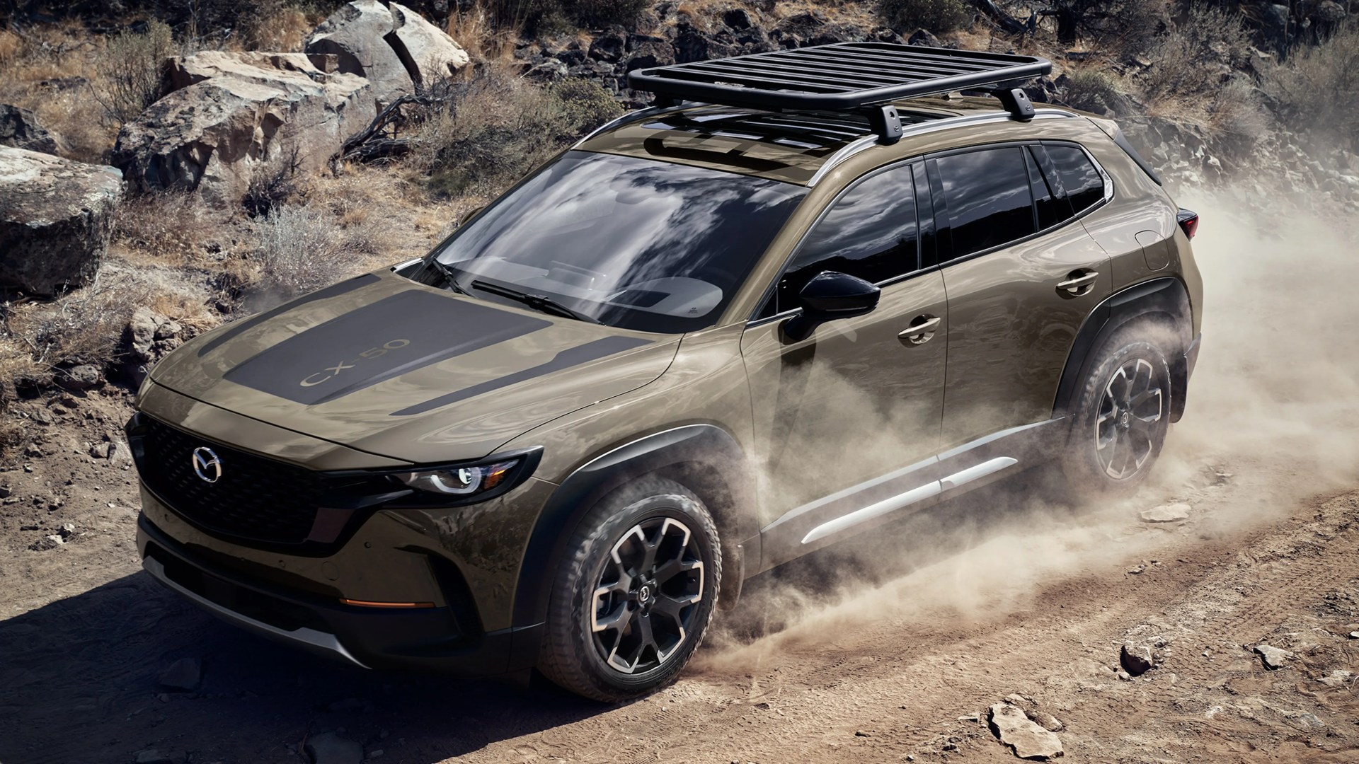 Olive green Mazda CX-50 SUV with roof rack driving on dusty rocky terrain.