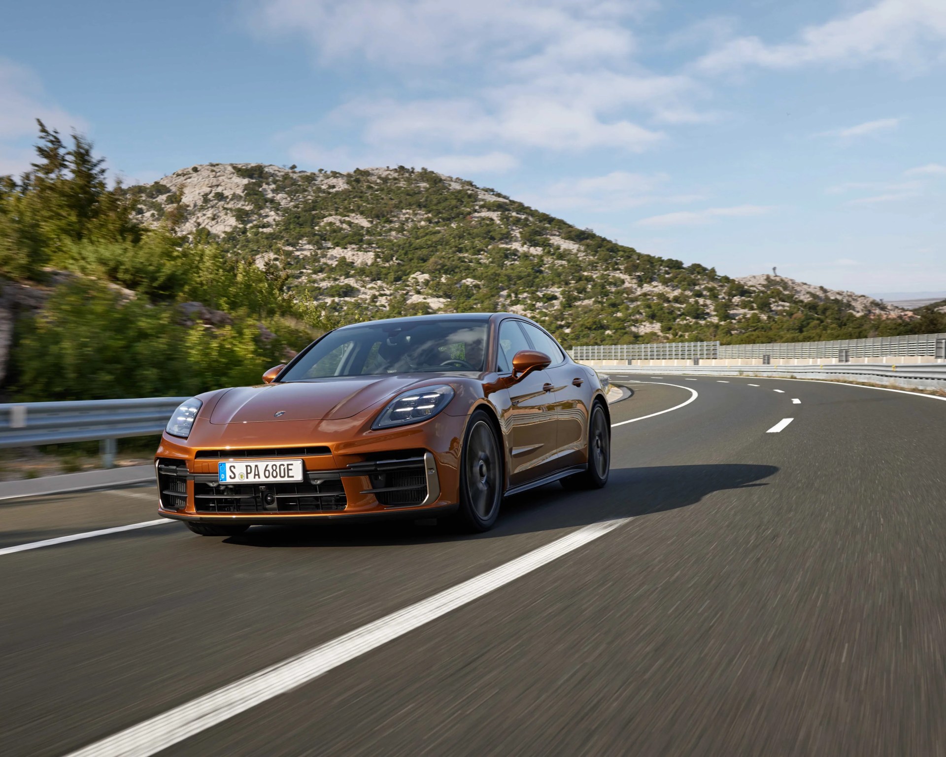 Metallic brown Porsche sedan driving on a curved mountain road under a partly cloudy sky.
