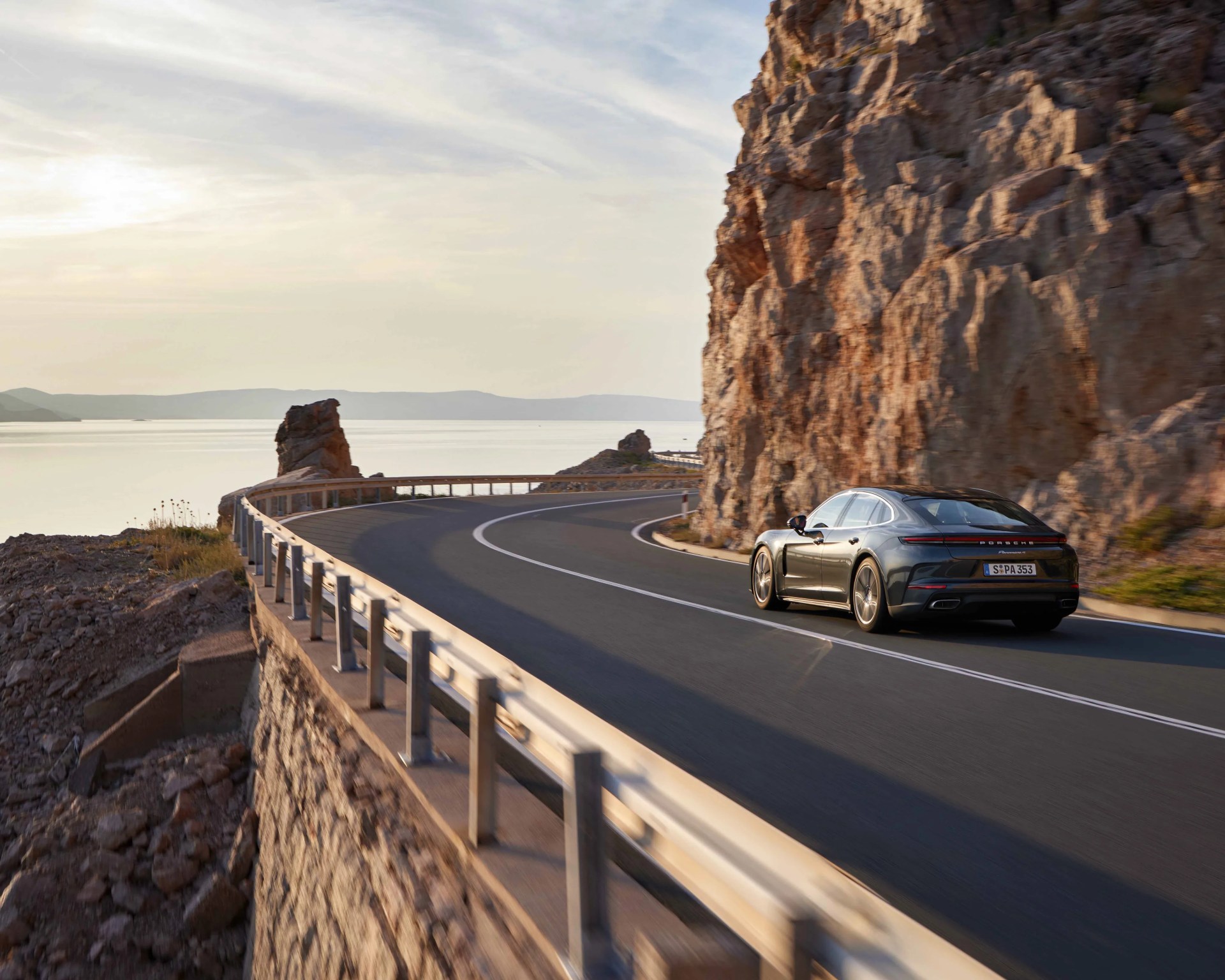 Black Porsche sedan driving on a coastal road with rocky cliffs and calm water at sunset.