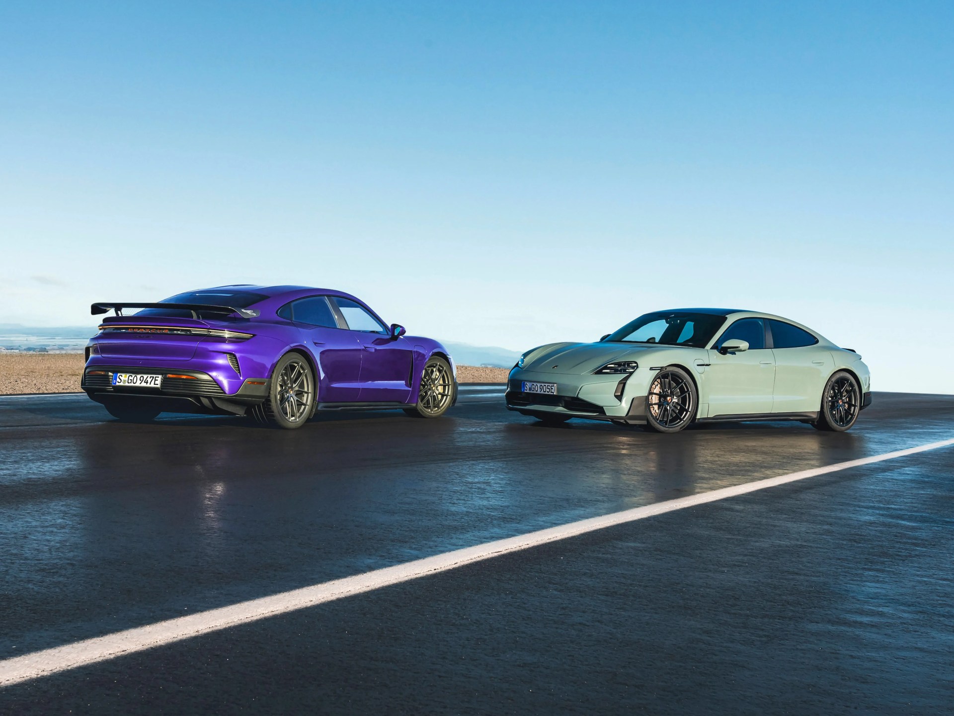 Two Porsche Taycan electric sports sedans, one purple with a rear spoiler and one light green, parked on a wet asphalt surface under a clear blue sky.