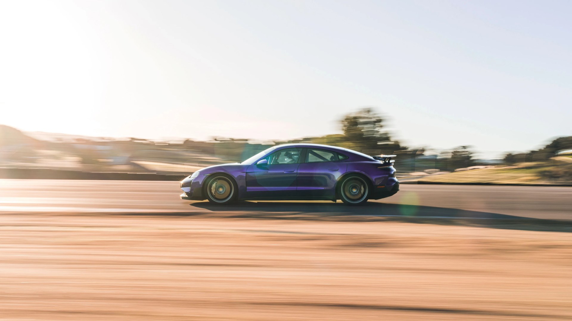 Purple sports sedan with a rear spoiler driving fast on a sunlit road.