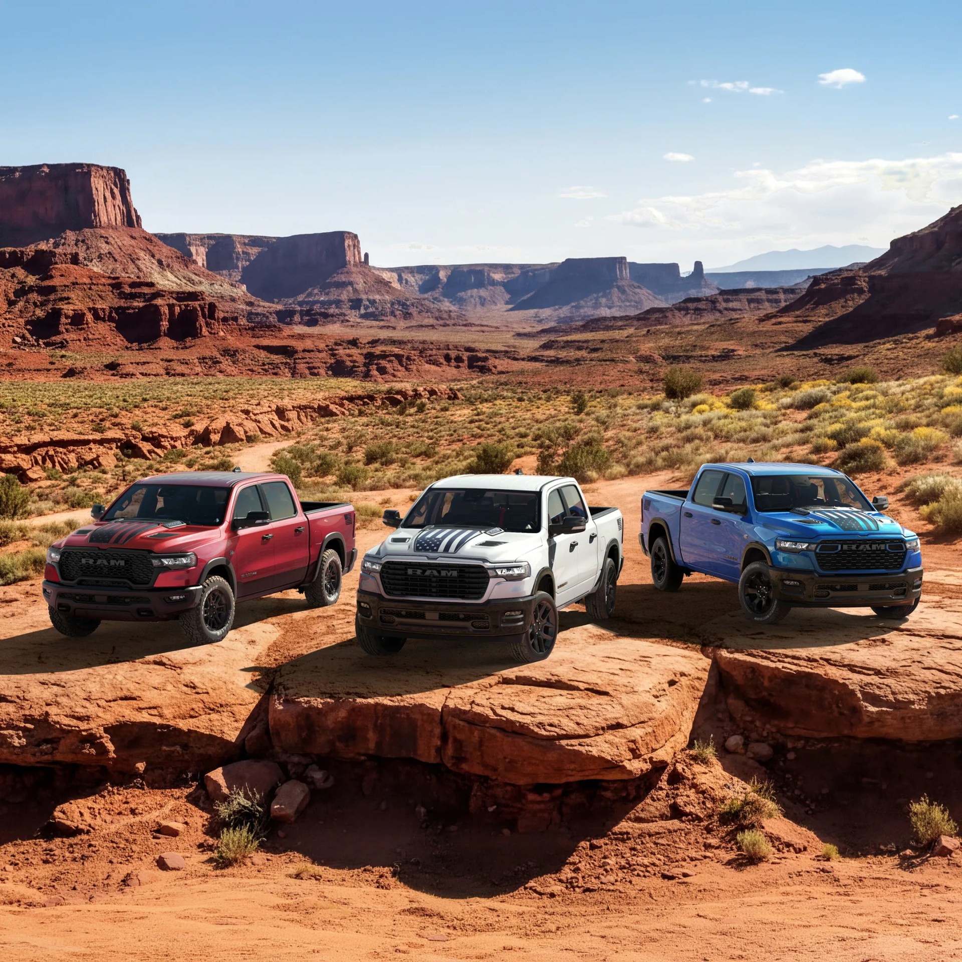 Three Ram pickup trucks in red, white, and blue with black racing stripes parked on rocky desert terrain with mesas in the background.