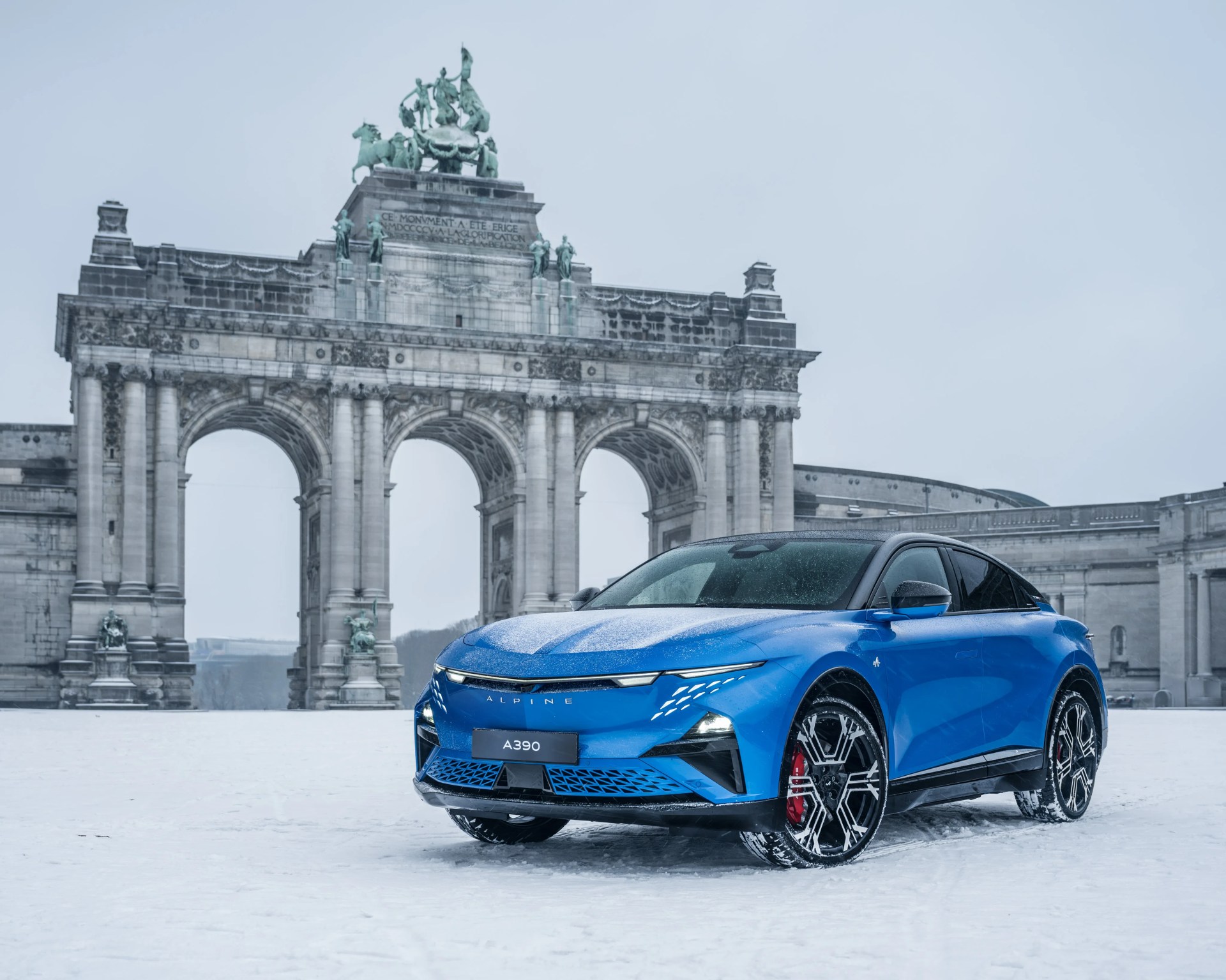 Blue Alpine A390 electric SUV with black roof and red brake calipers parked on snow in front of a stone arch monument.
