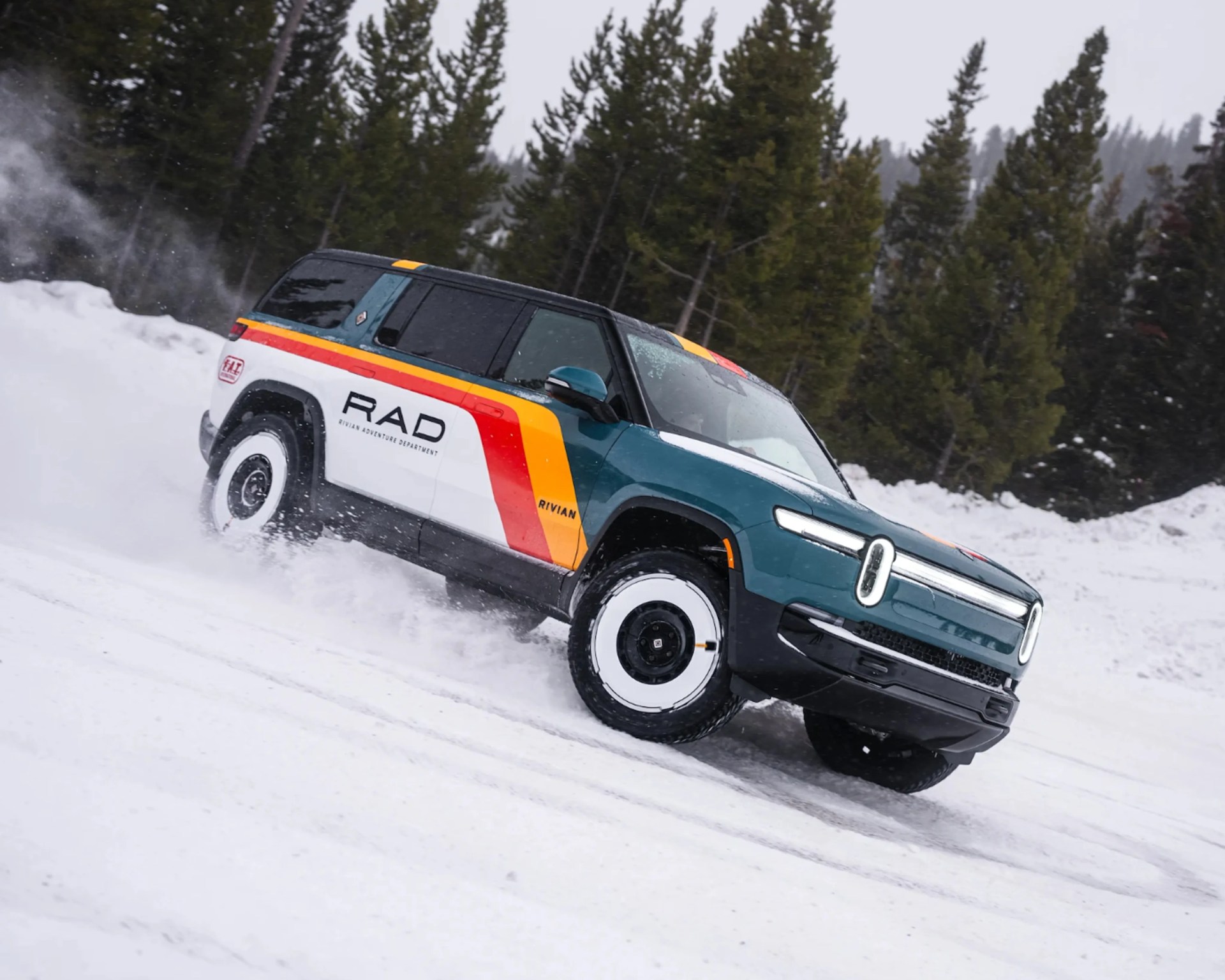 Teal and white Rivian SUV with RAD logo and orange-yellow stripes driving on snow near pine trees.