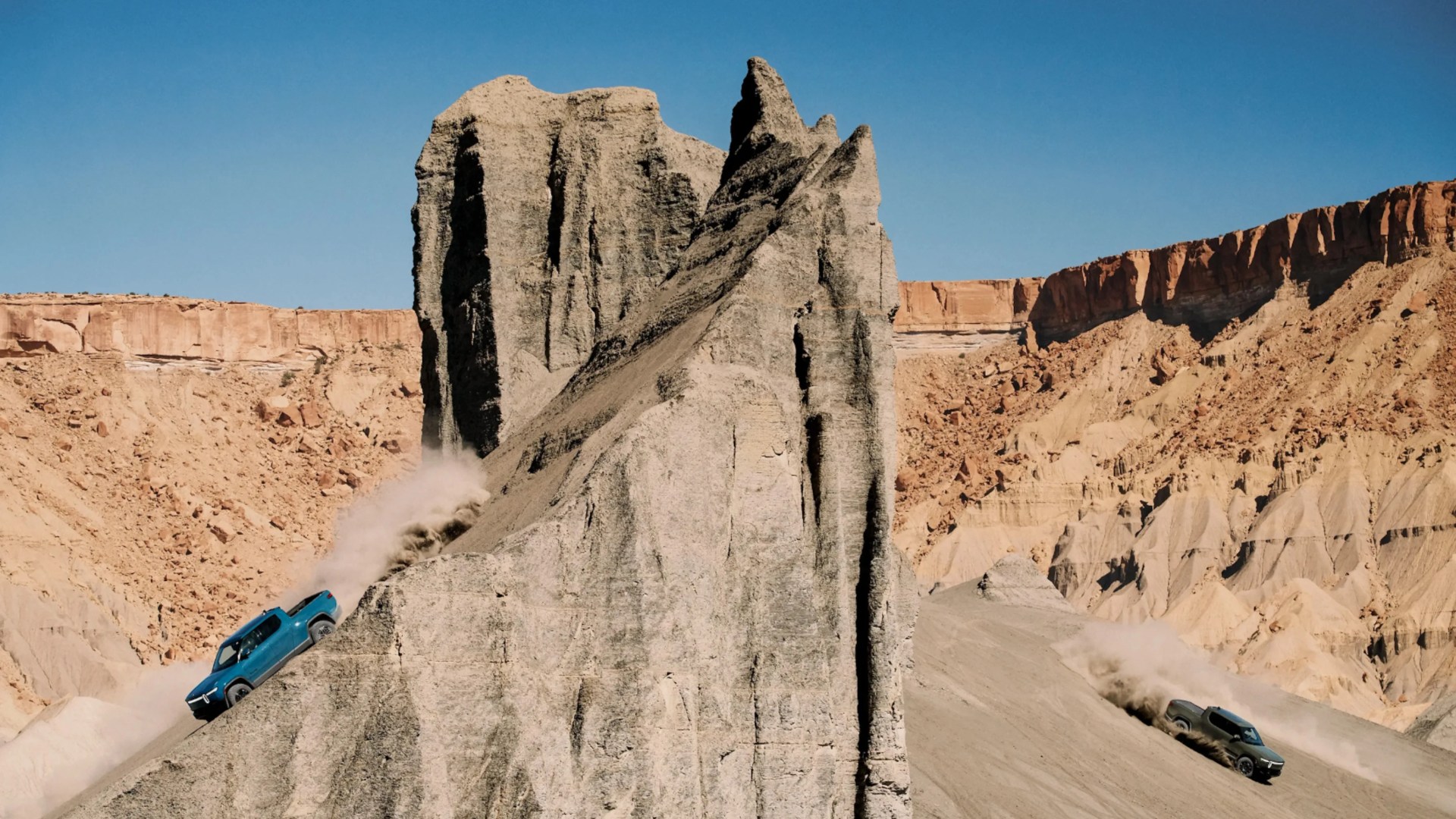 Two off-road vehicles kicking up dust while descending steep rocky slopes in a desert landscape.
