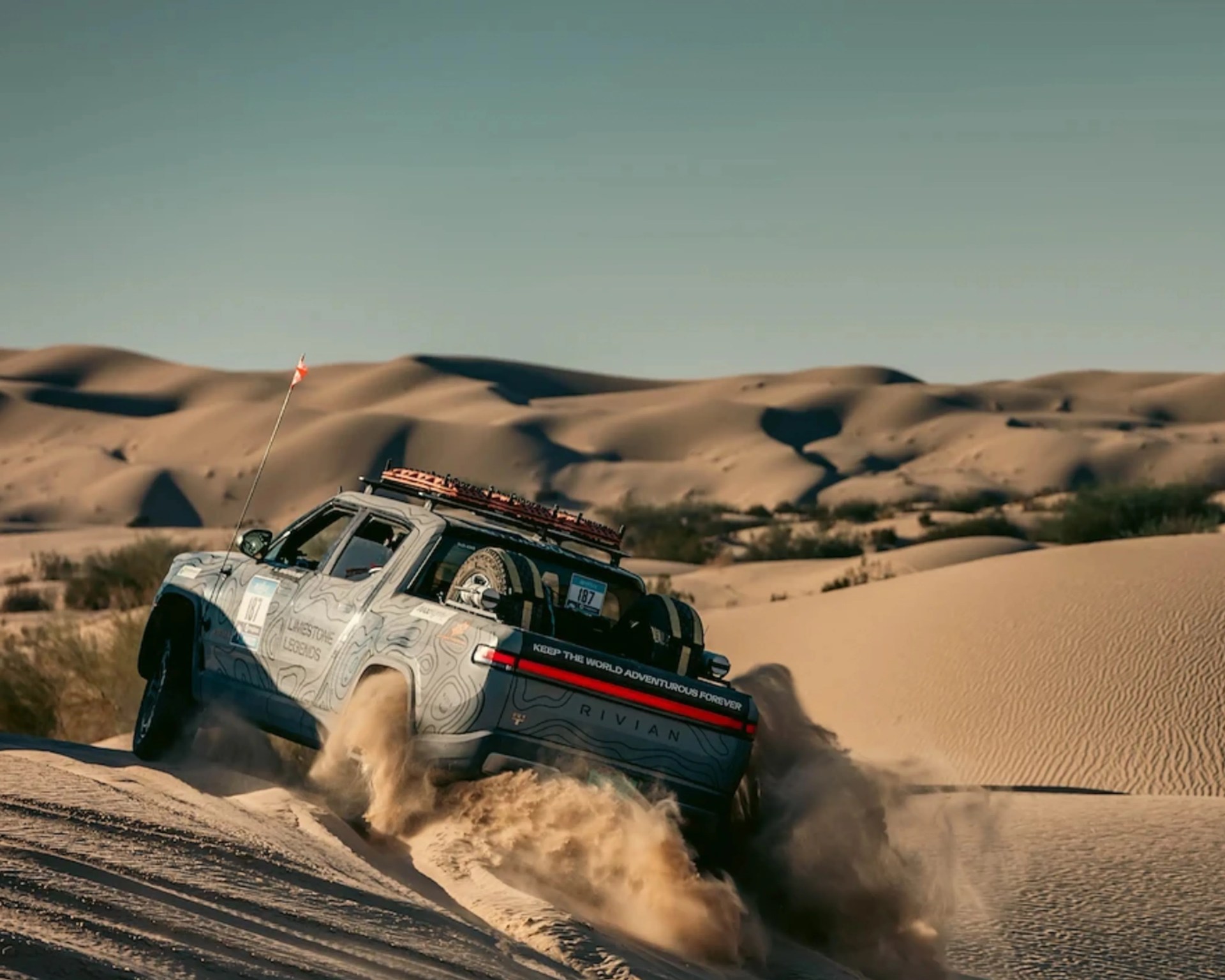 Rivian electric truck kicking up sand while driving uphill in a desert with sand dunes in the background.