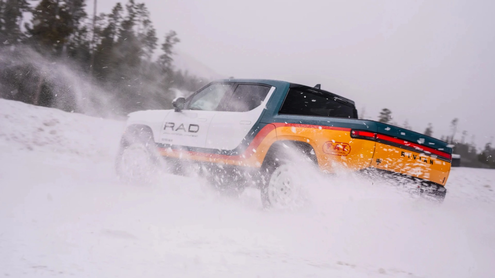 Multicolor Rivian pickup truck driving through snow with snow spray around the wheels.