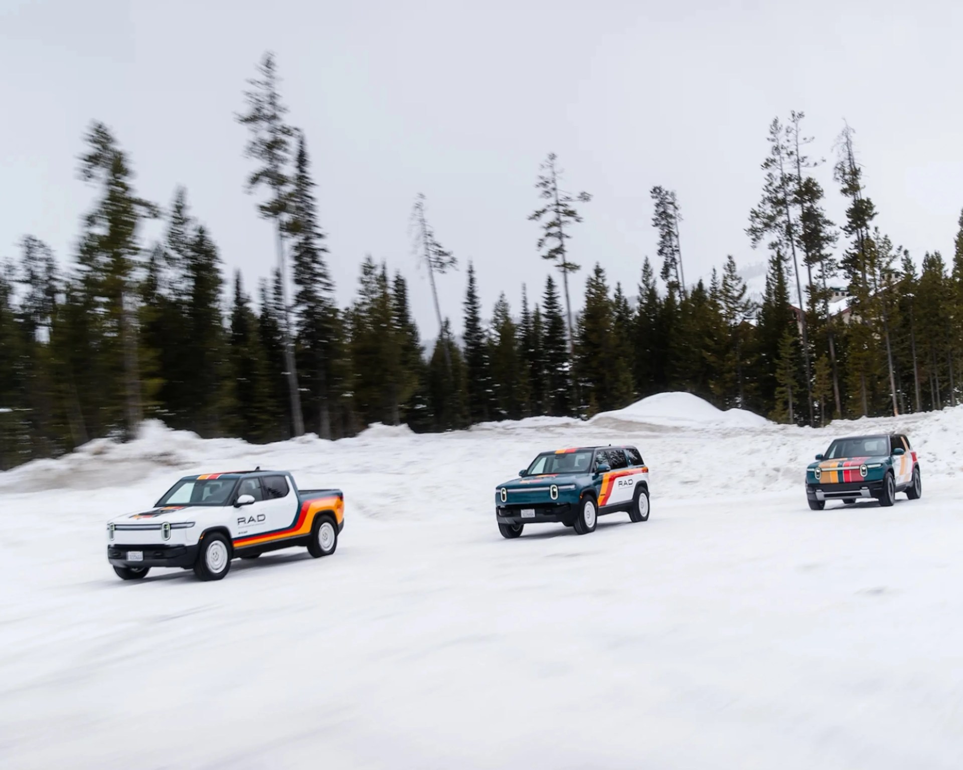 Three electric SUVs and a pickup truck with RAD branding and colorful stripes driving on a snowy road with pine trees in the background.