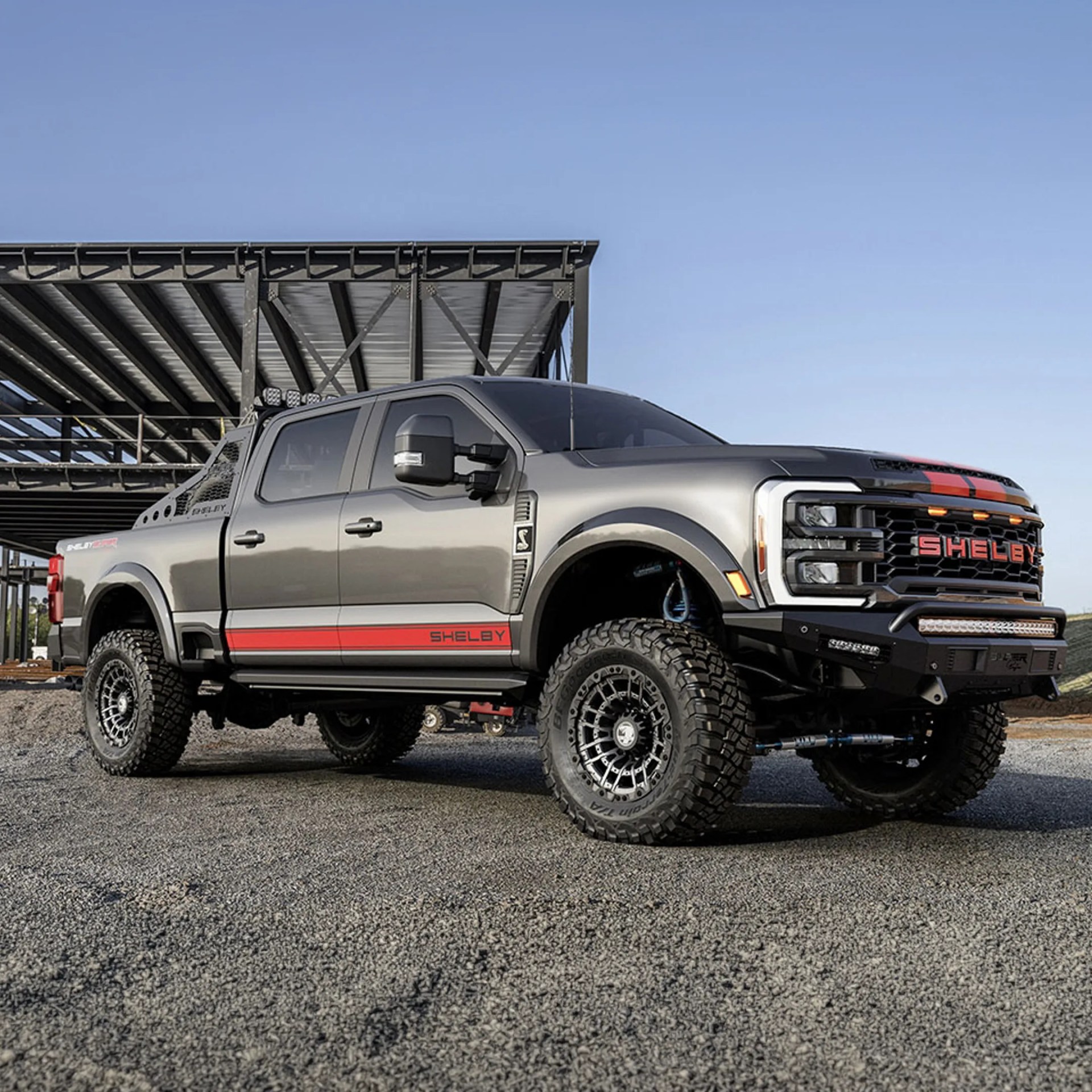 Gray Shelby off-road pickup truck with red racing stripes and large all-terrain tires parked on gravel.