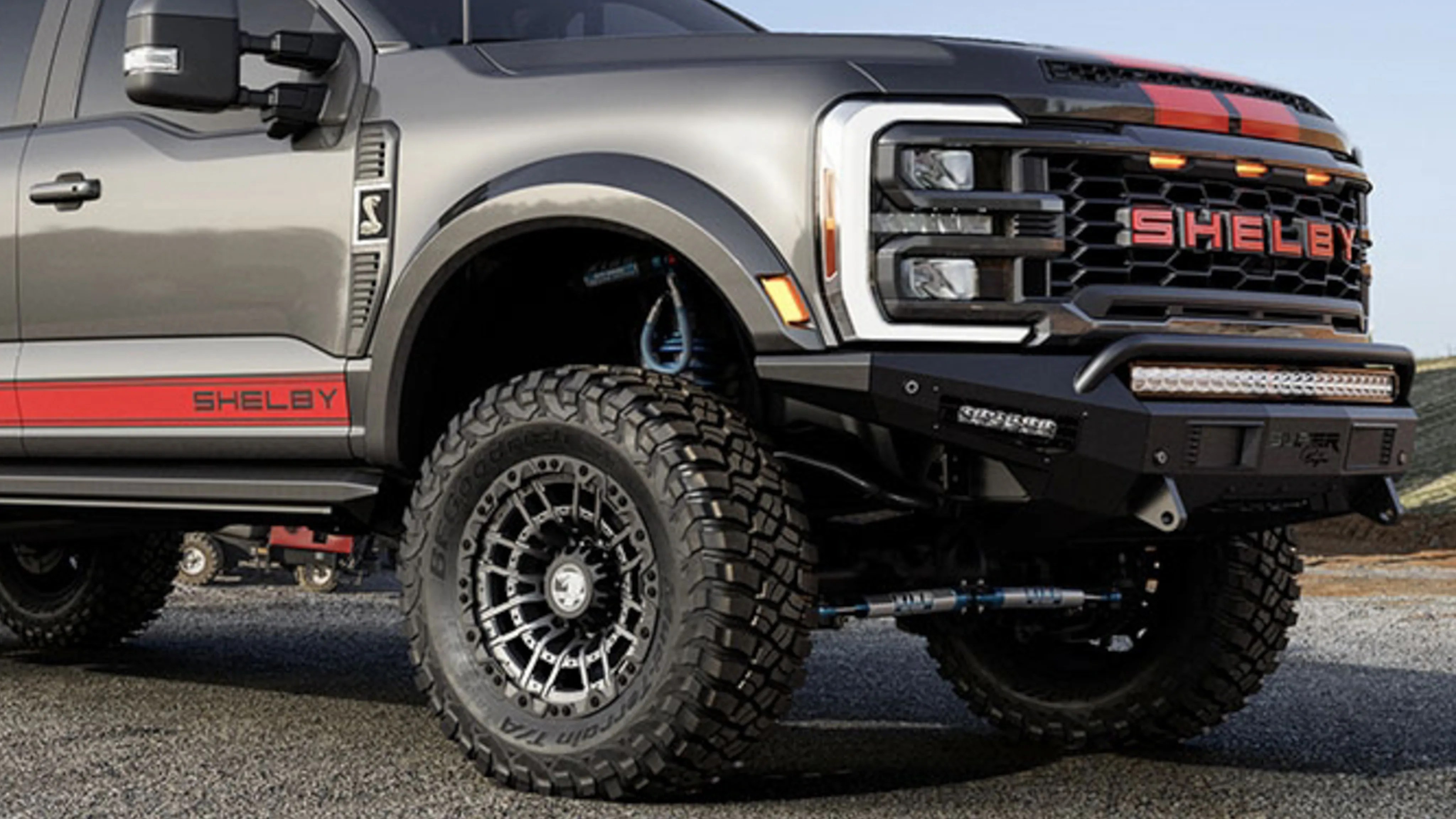 Gray Shelby truck front side with large off-road tires, red stripe and Shelby logo on the grille and side panel.