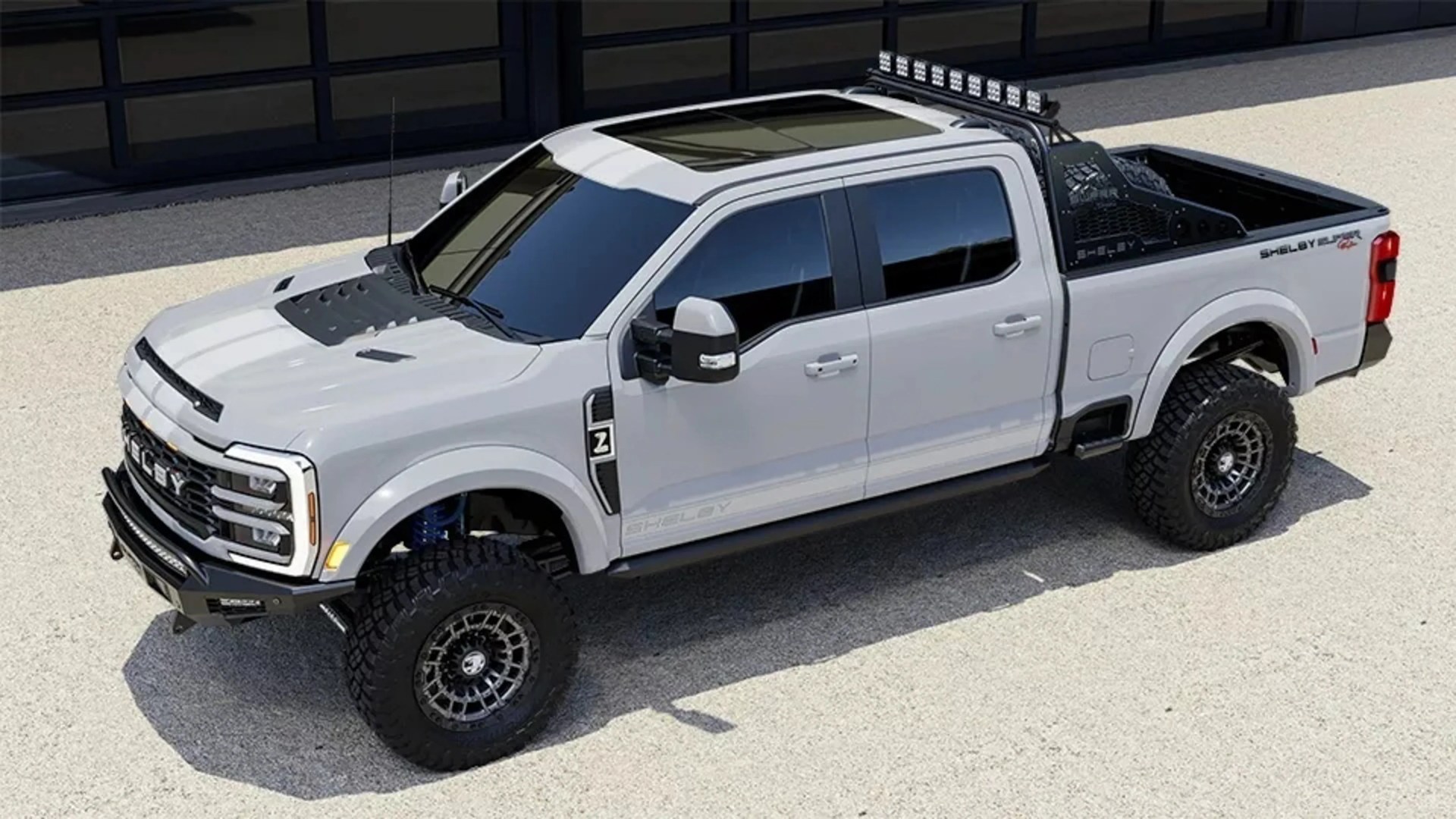 White Shelby Super Crew pickup truck with black off-road tires and roof light bar parked on gravel.