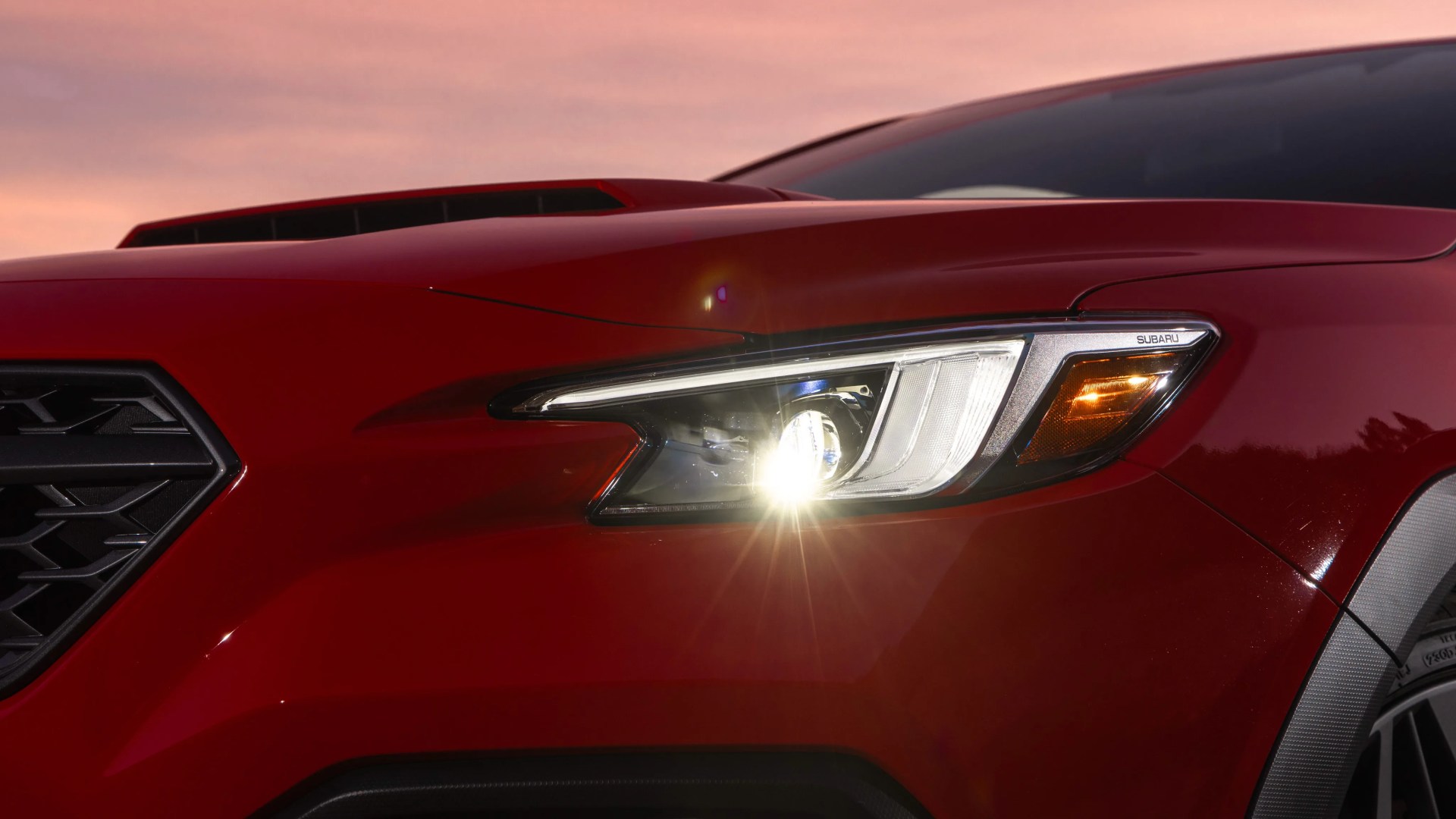 Close-up of a red Subaru car's front headlight with the light turned on at sunset.