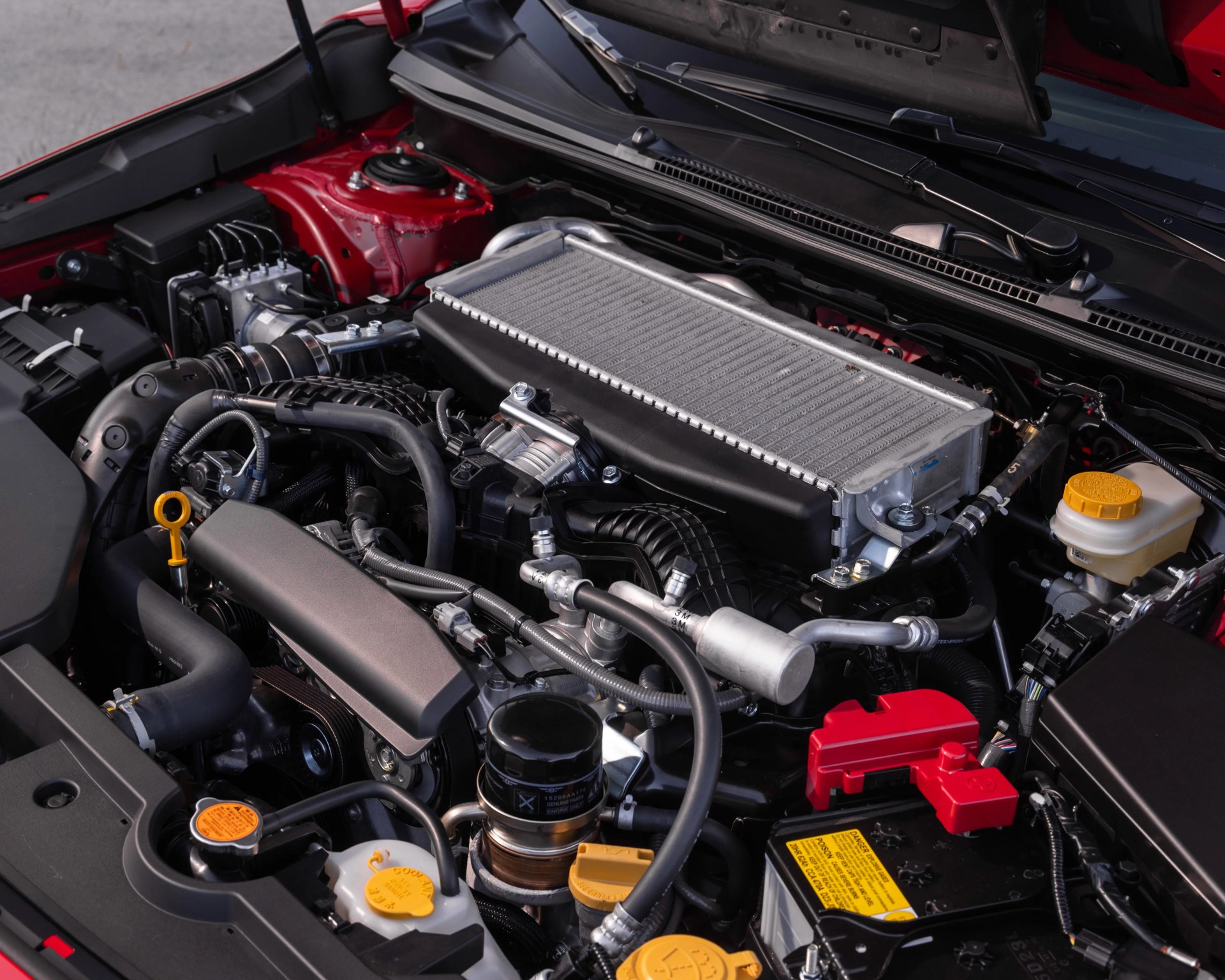 Car engine bay with visible intercooler, battery with red cover, and various hoses and fluid reservoirs.