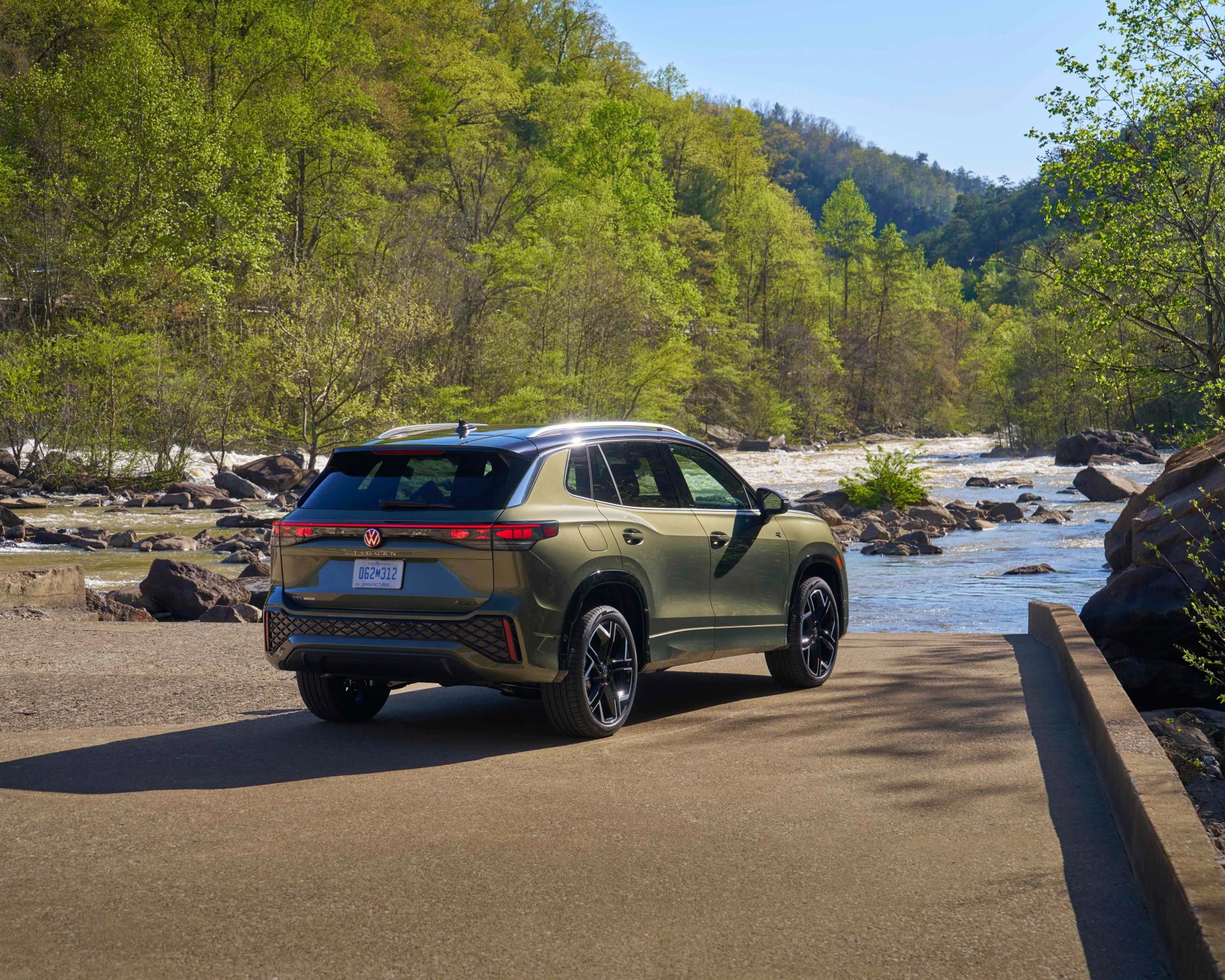 Olive green Volkswagen SUV parked on a concrete surface near a river with green trees and hills in the background.