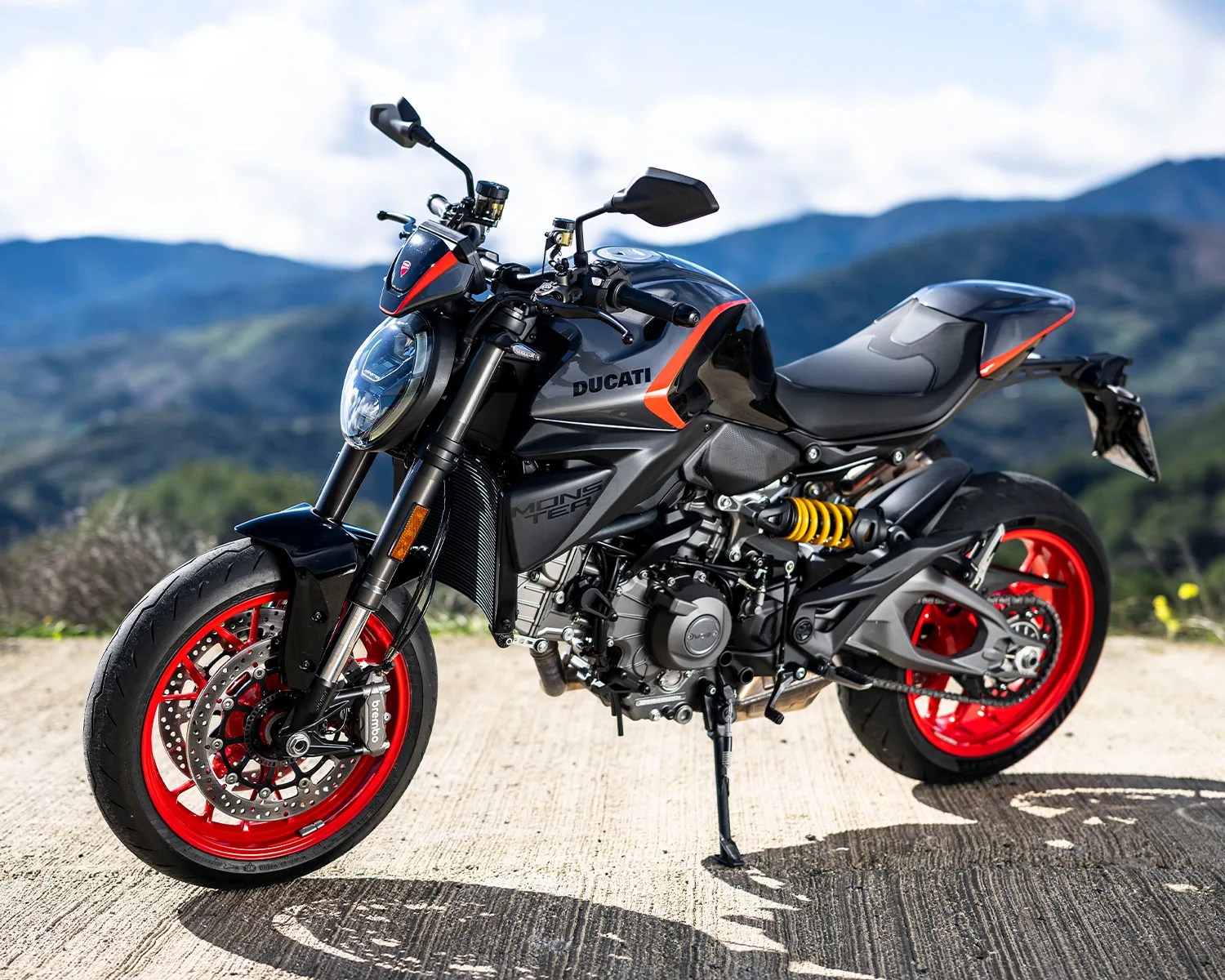 Black and gray Ducati Monster motorcycle with red wheels and yellow suspension parked on a concrete surface with mountains in the background.