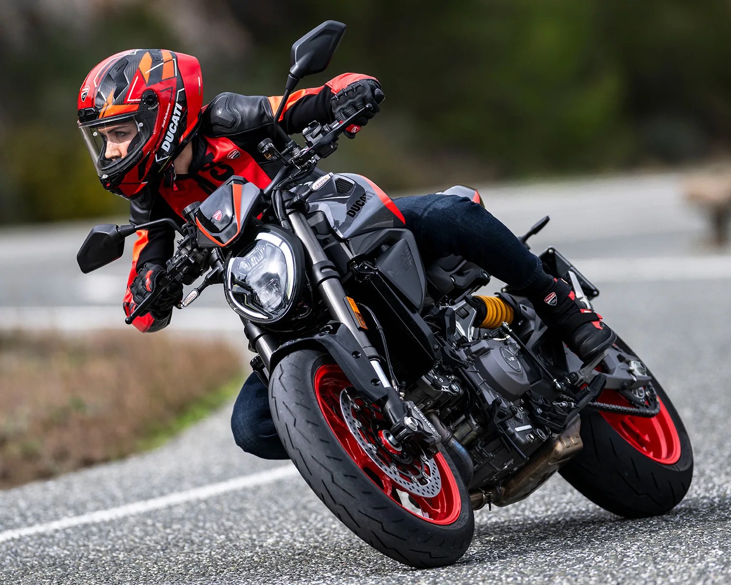 Person wearing a red and black Ducati helmet and jacket riding a gray Ducati motorcycle with red rims on a road.