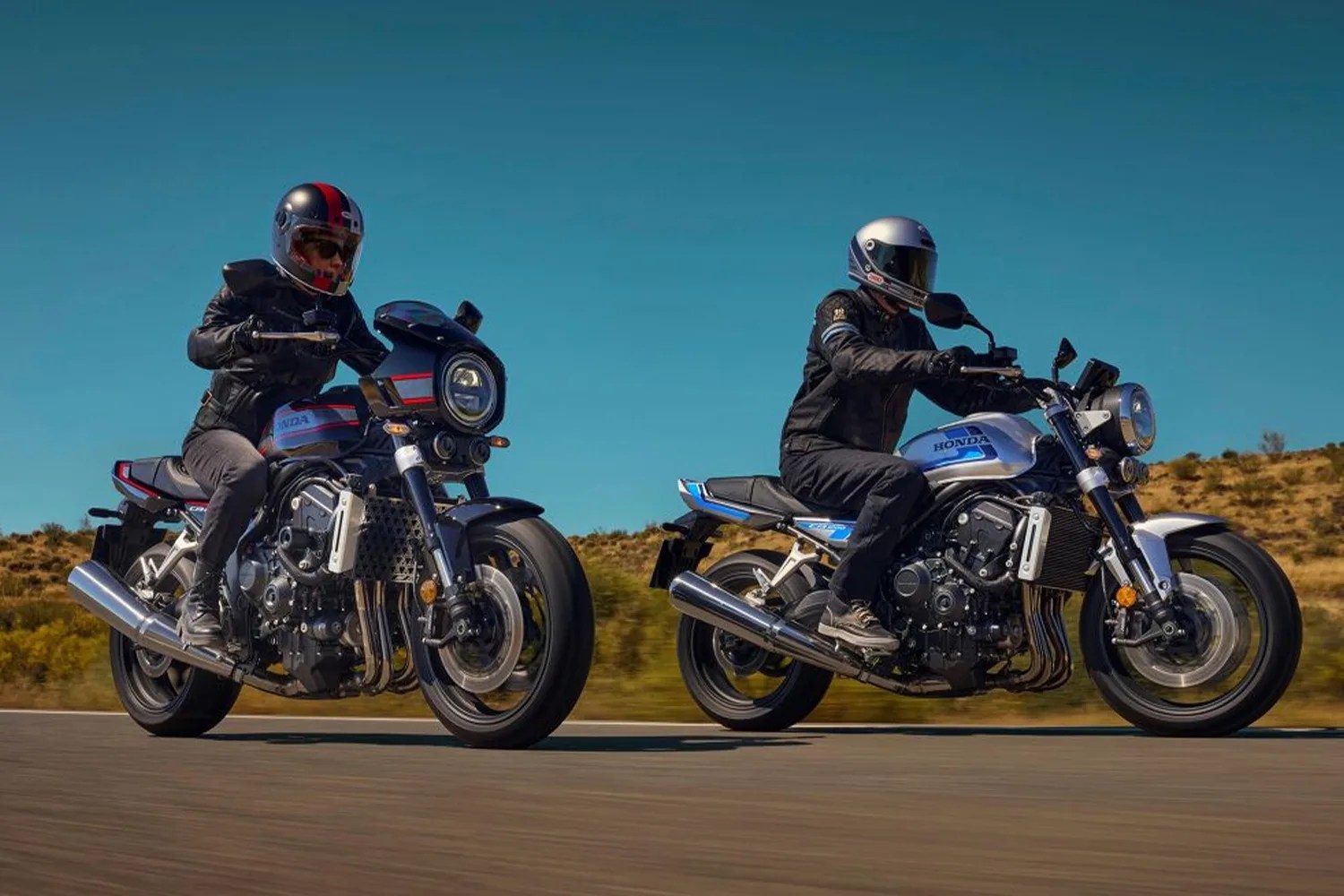 Two riders wearing helmets and black jackets on silver and black Honda motorcycles on a road with clear blue sky.
