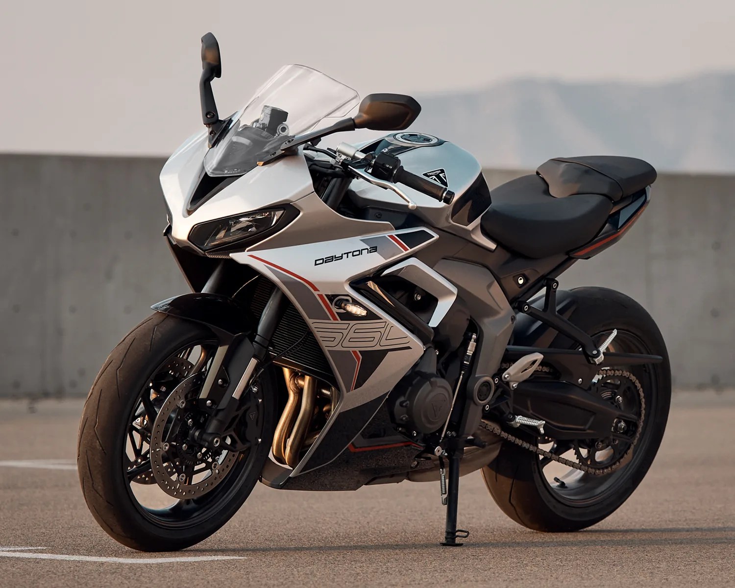 Silver and black Triumph Daytona 765 sport motorcycle parked on asphalt with a concrete wall in the background.