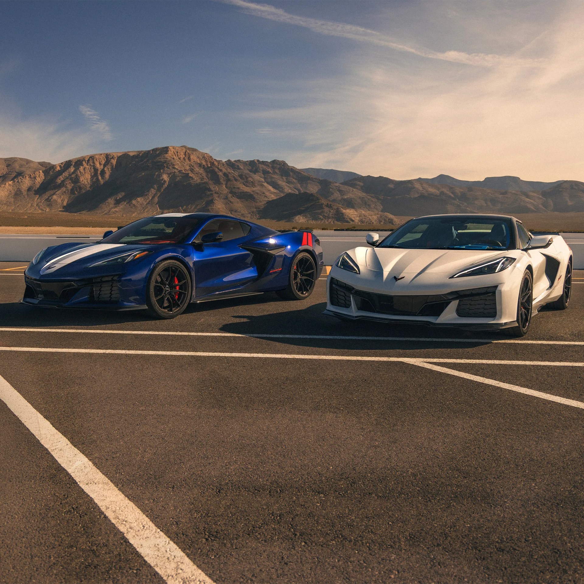Blue sports car with white racing stripes and red accents parked next to a white sports car on an asphalt lot with mountains in the background.