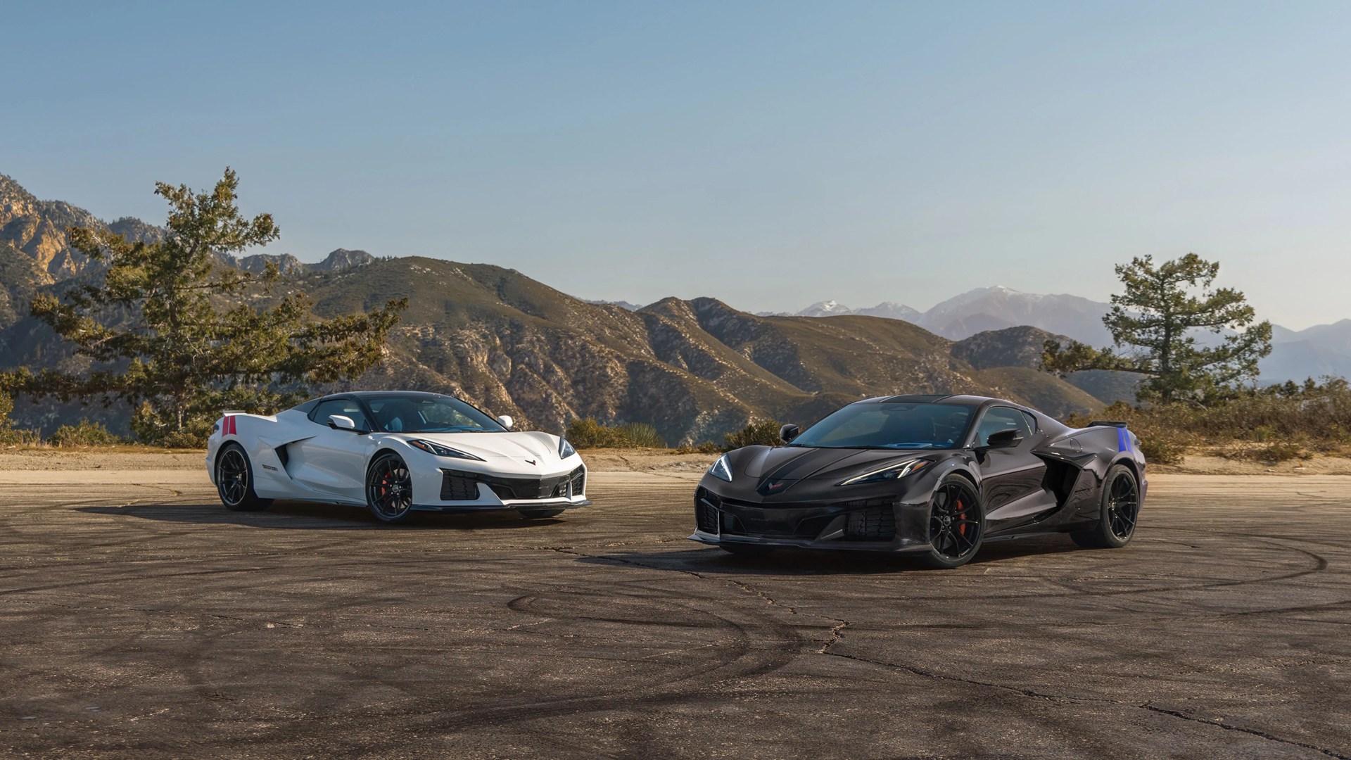 Two Chevrolet Corvette sports cars, one white and one black, parked on an asphalt surface with mountains and trees in the background.