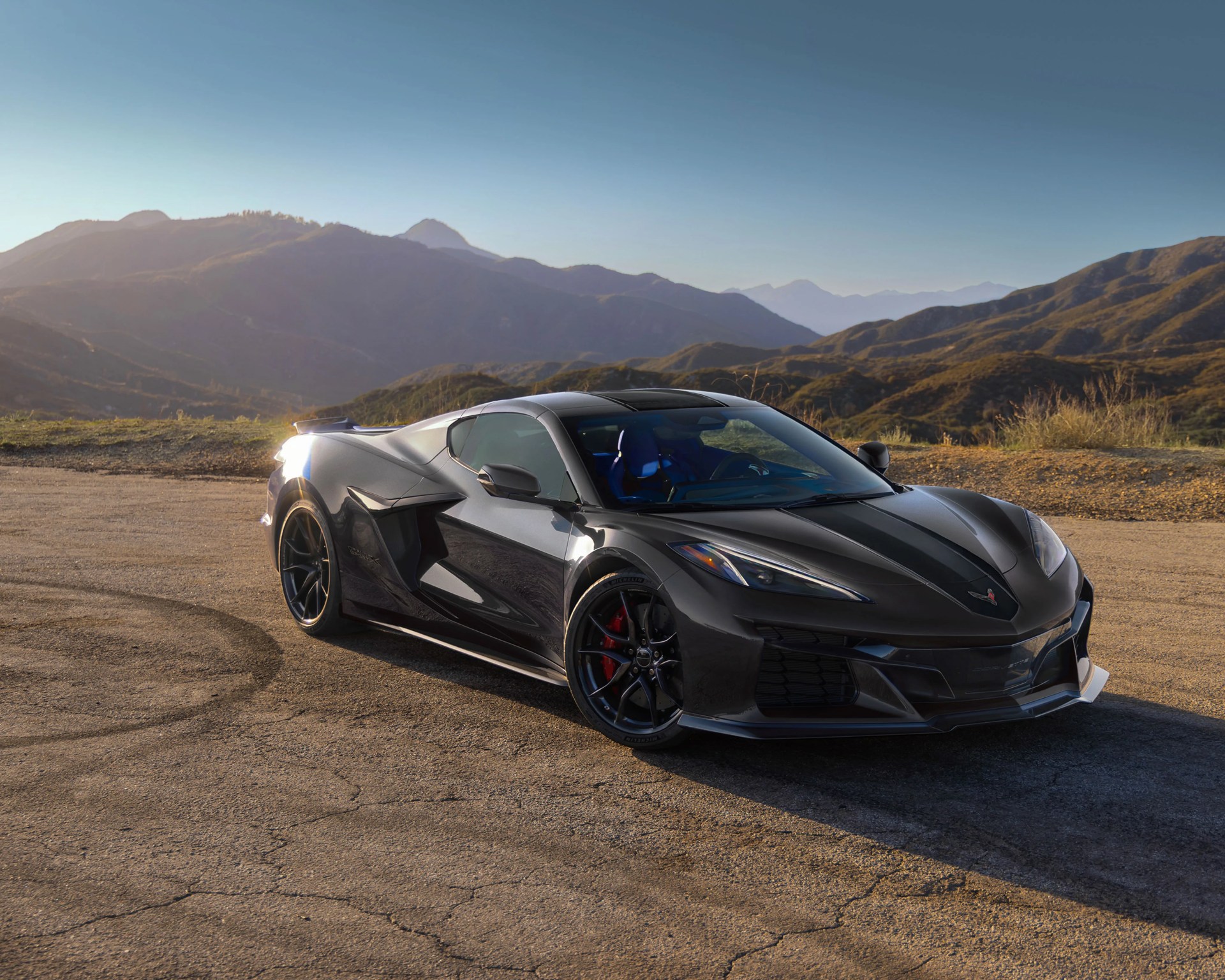 Glossy black sports car with blue interior parked on cracked pavement with mountains in the background.