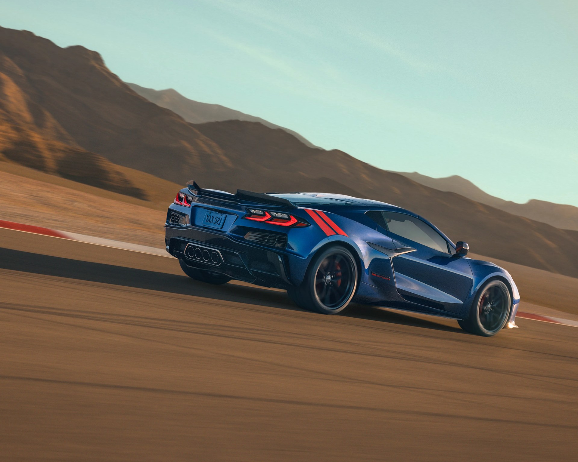 Blue sports car with red racing stripes driving on a desert race track with mountains in the background.
