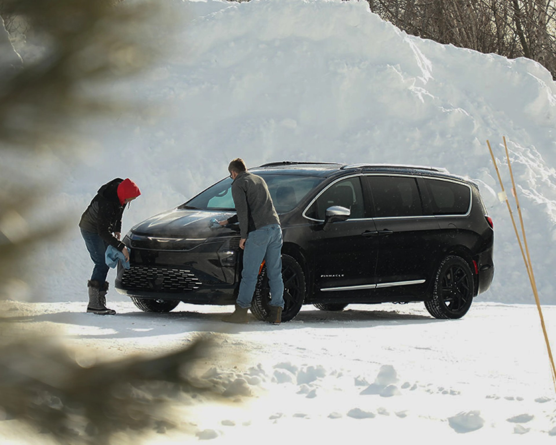 Two people cleaning a black Chrysler Pacifica minivan in a snowy outdoor setting.