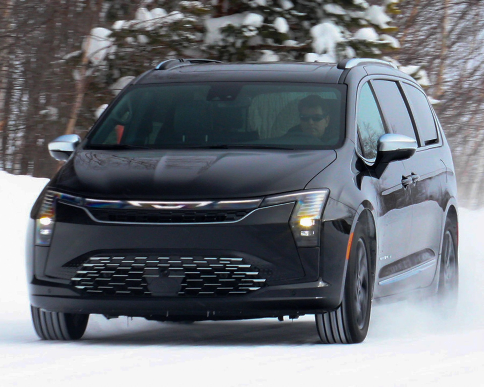 Black minivan with modern LED headlights driving on snow with trees in the background.