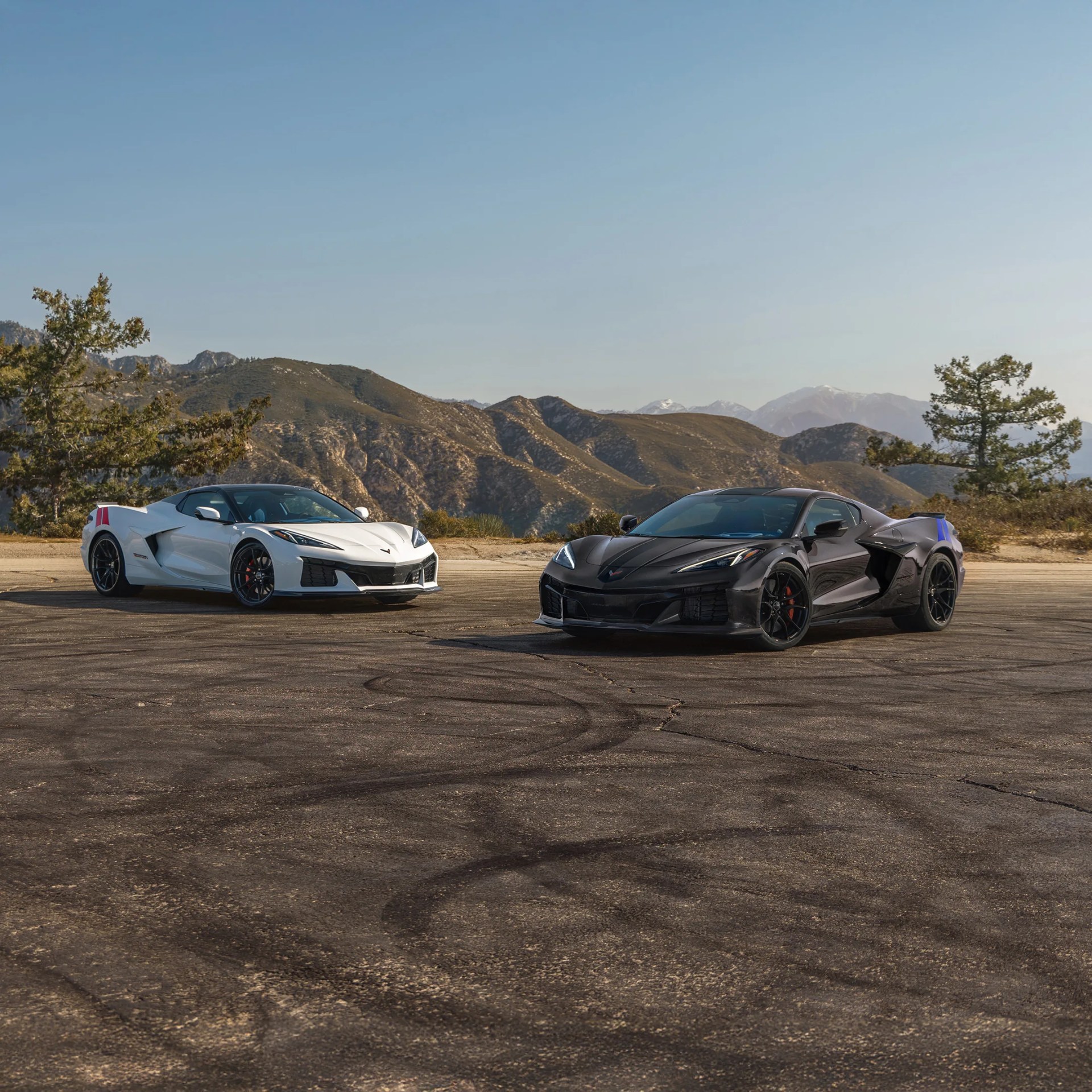 Two Chevrolet Corvette sports cars, one white and one black, parked on an asphalt surface with mountains and trees in the background.