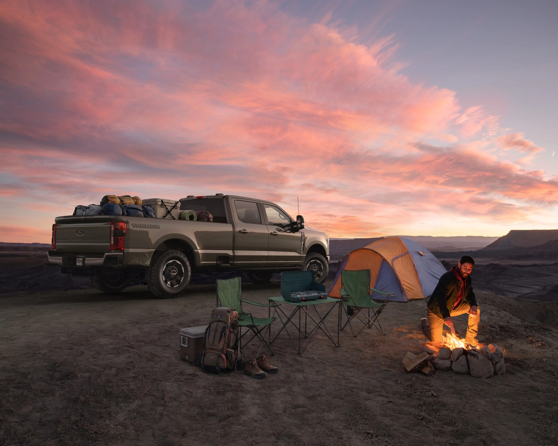 Tan Ford Super Duty Tremor pickup truck loaded with camping gear next to a blue and orange tent, green folding chairs, and a man tending a campfire at sunset.