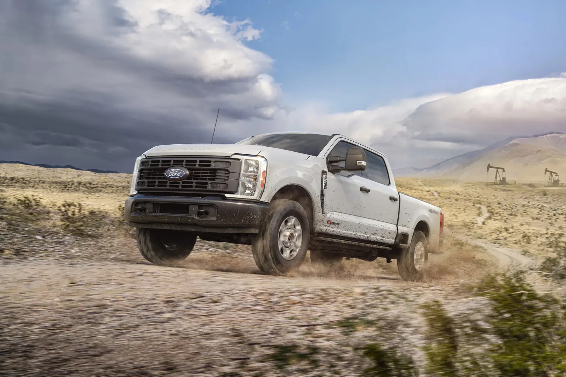 White Ford Super Duty pickup truck driving off-road on a dusty desert trail with oil pump jacks in the background.