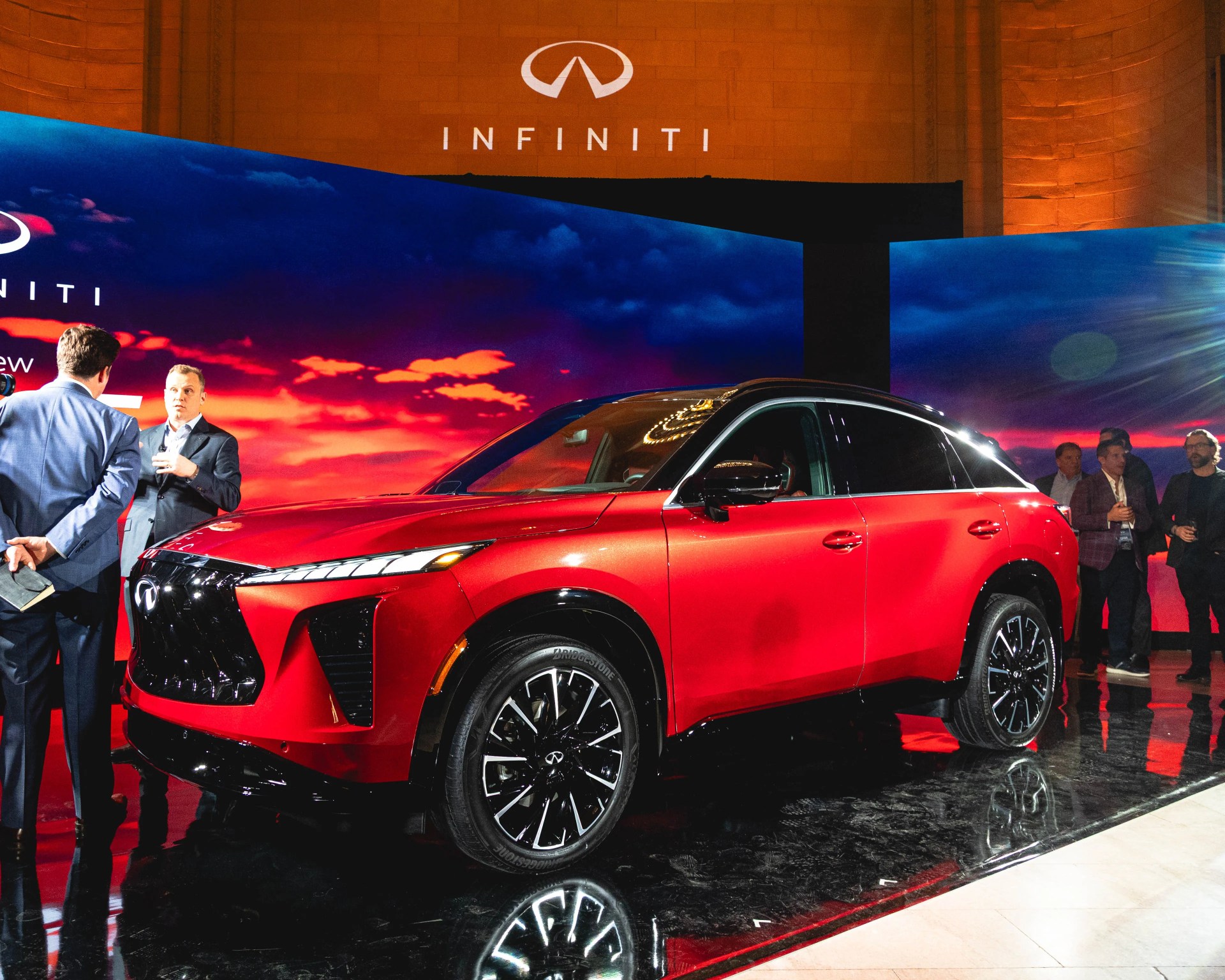 Red Infiniti SUV with black roof and alloy wheels displayed indoors with people and Infiniti logo in background.