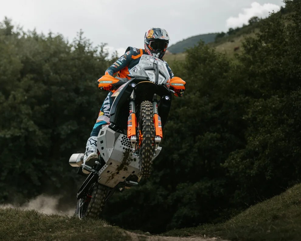 Off-road motorcycle with orange and black accents jumping on a dirt trail with trees in the background.