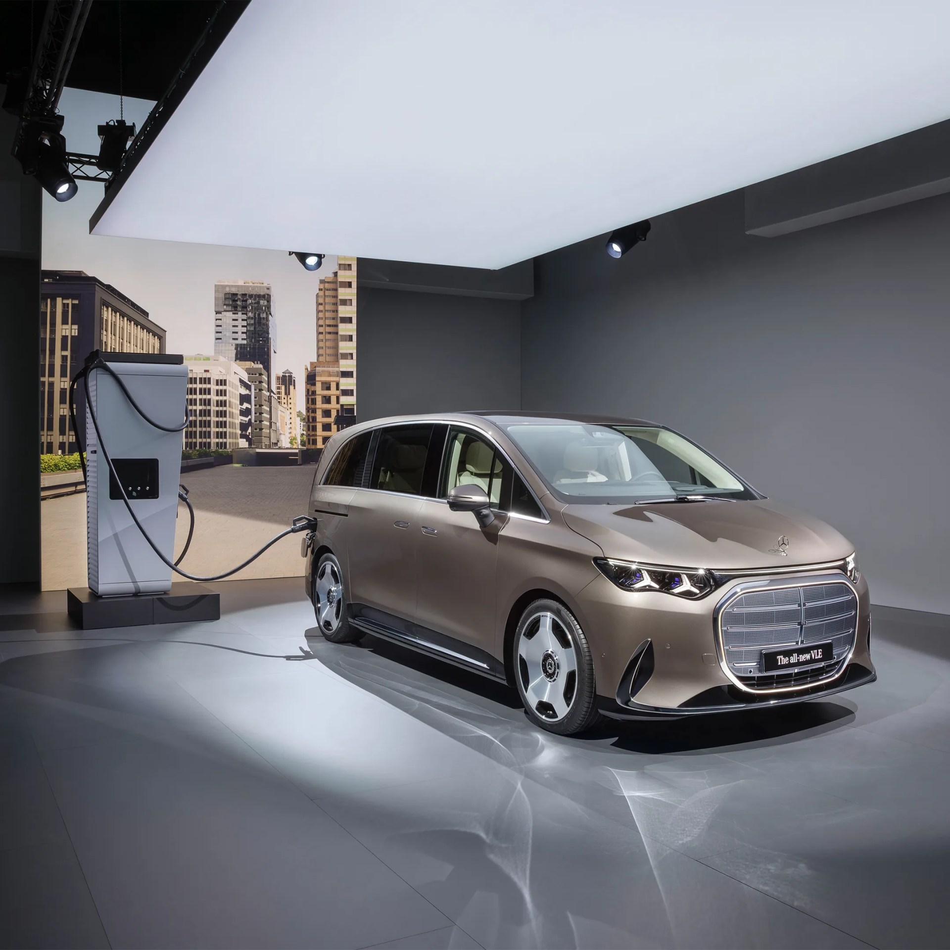 Metallic brown Mercedes-Benz electric minivan charging at a station indoors with cityscape backdrop.