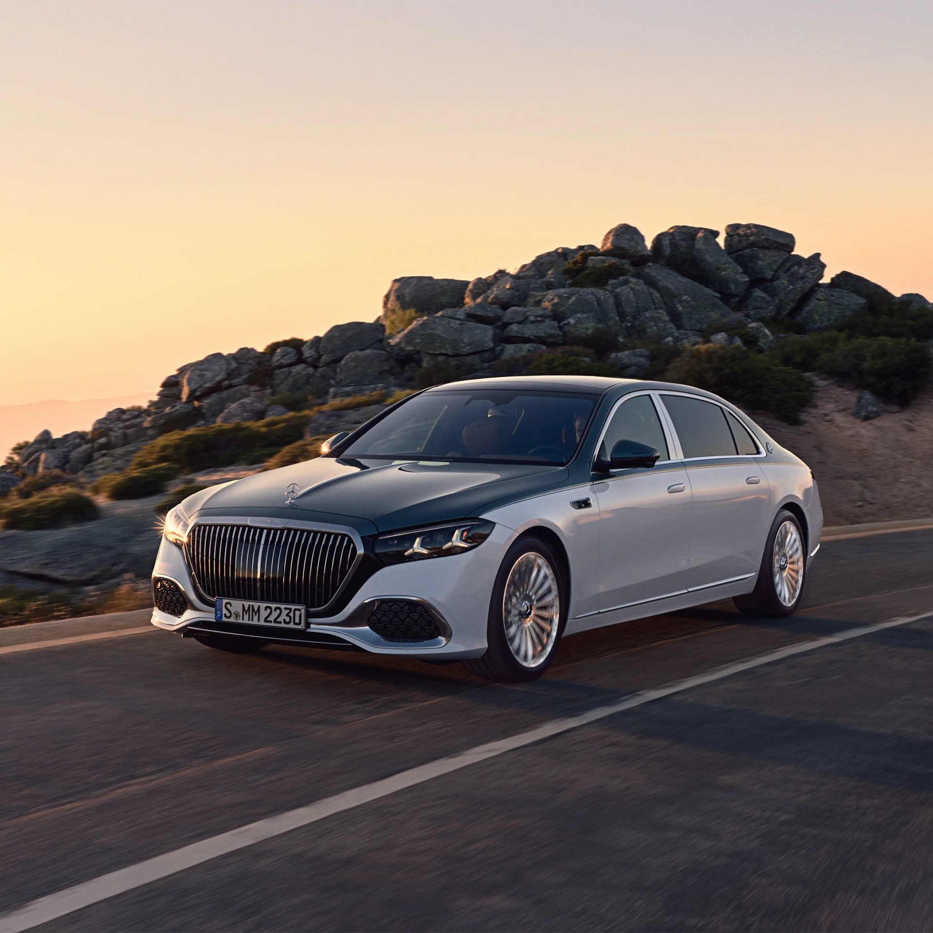 Two-tone silver and black luxury sedan with large vertical grille driving on a mountain road at sunset.