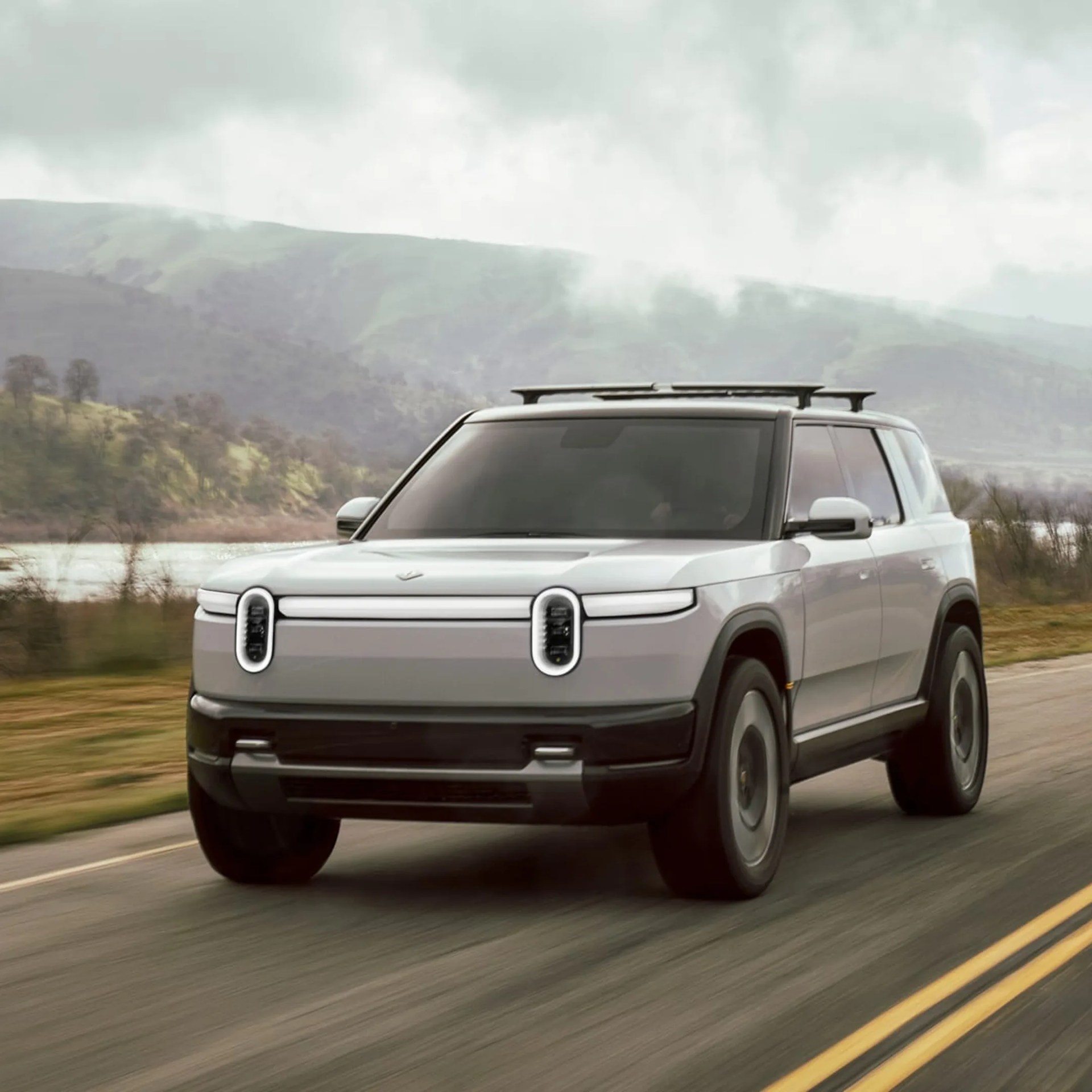 Silver Rivian R1S electric SUV driving on a road with mountains and a lake in the background.