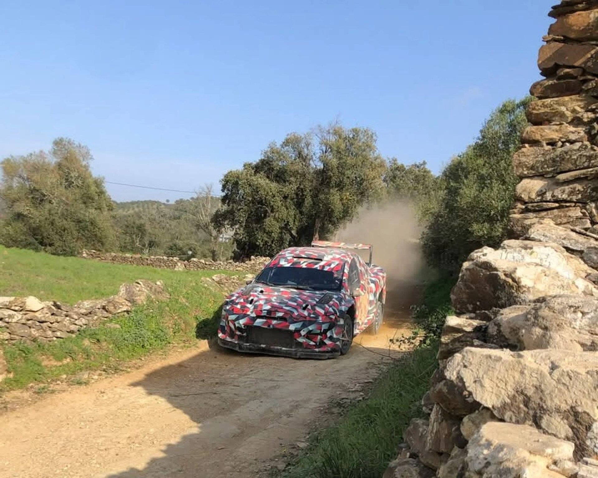 Rally car with red, black, and white camouflage driving on a dirt road with dust behind it.