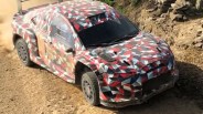 Rally car with red, black, white, and gray geometric camouflage driving on a dirt road.