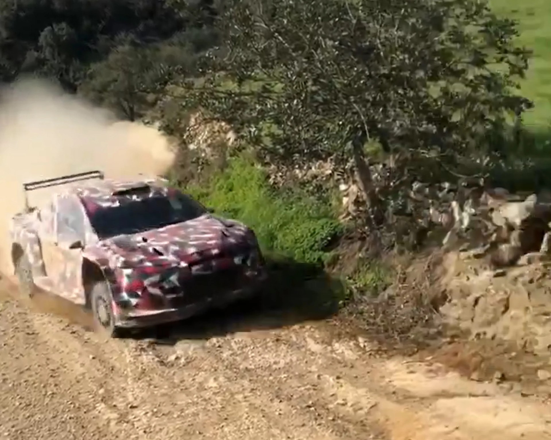 Rally car with camouflage paint kicking up dust on a dirt road near greenery and rocks.